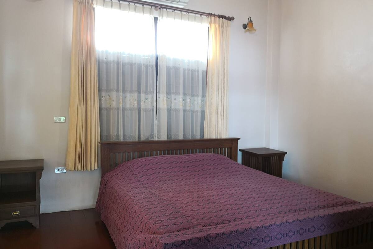 A bedroom with a bed, a nightstand, a wooden dresser, a window with curtains, and an air conditioning unit.