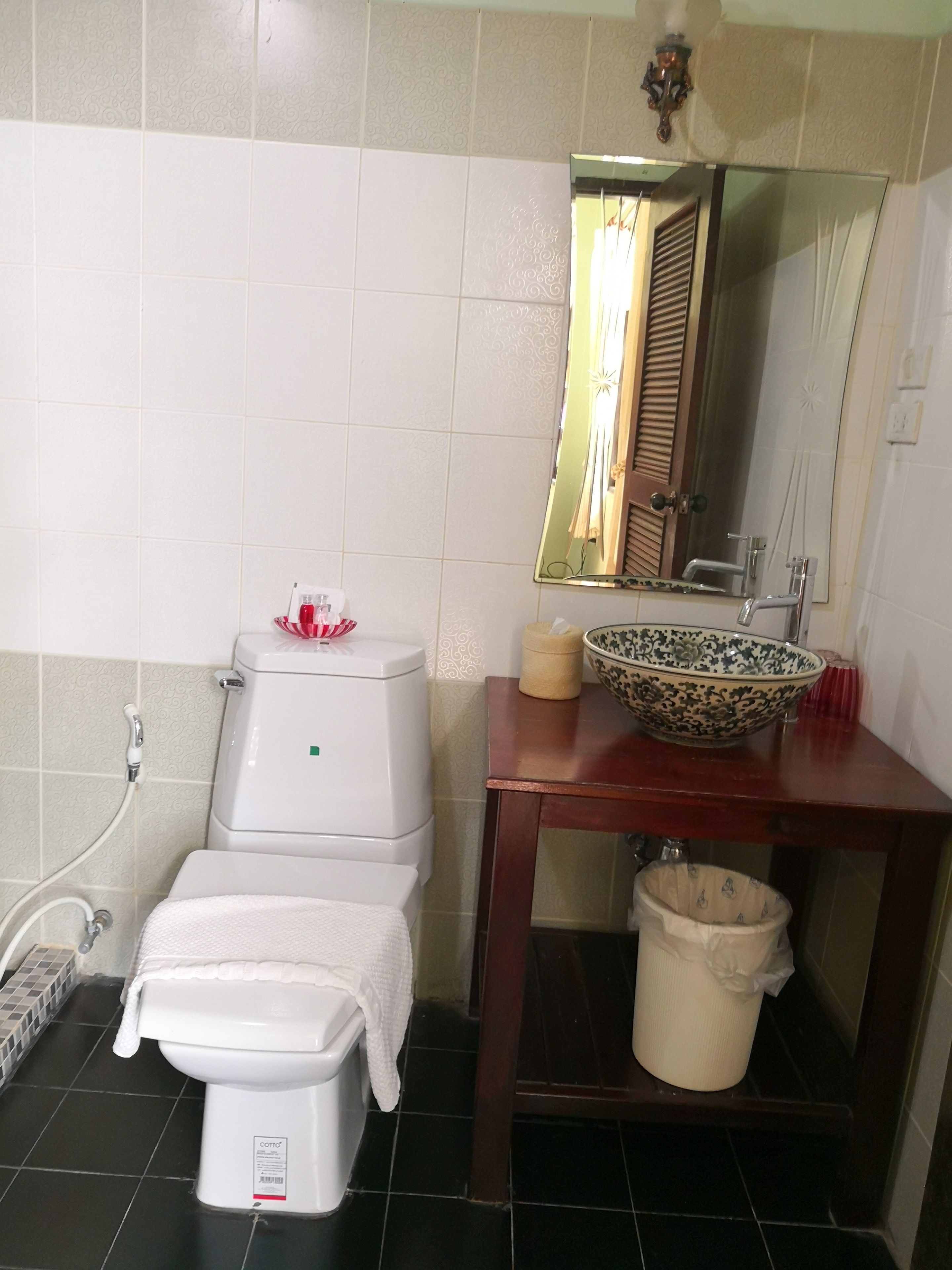 Un baño con inodoro, lavamanos con un recipiente decorativo, espejo y cubo de basura.