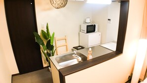 A compact hotel room with a dark brown vanity, a white sink, a microwave, a small refrigerator, a wooden chair, and a potted plant. - One More Heart at Kamishichiken 2 (Kyoto)