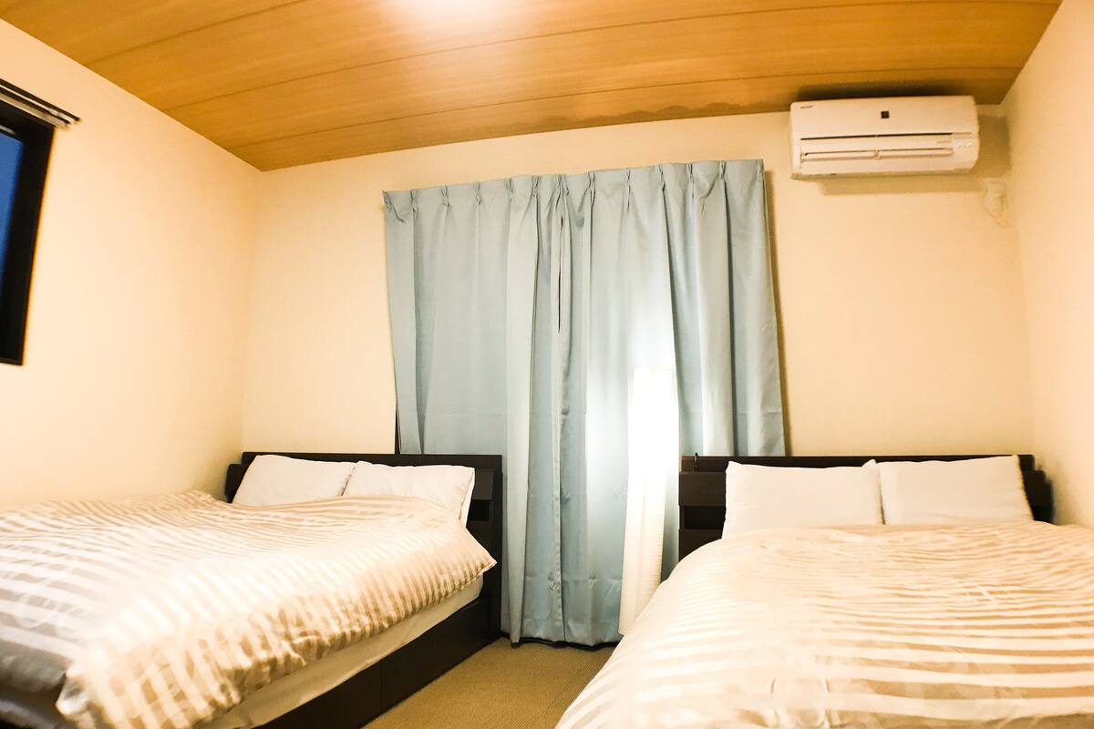 A room with two beds, a window with curtains, an air conditioning unit, and a wooden ceiling.