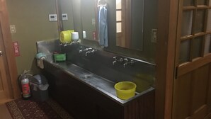 Ein Badezimmer mit einem großen Waschbecken, einem Spiegel und einem Handtuchhalter. Ein Mülleimer und eine Flasche Handdesinfektionsmittel stehen auf dem Boden.