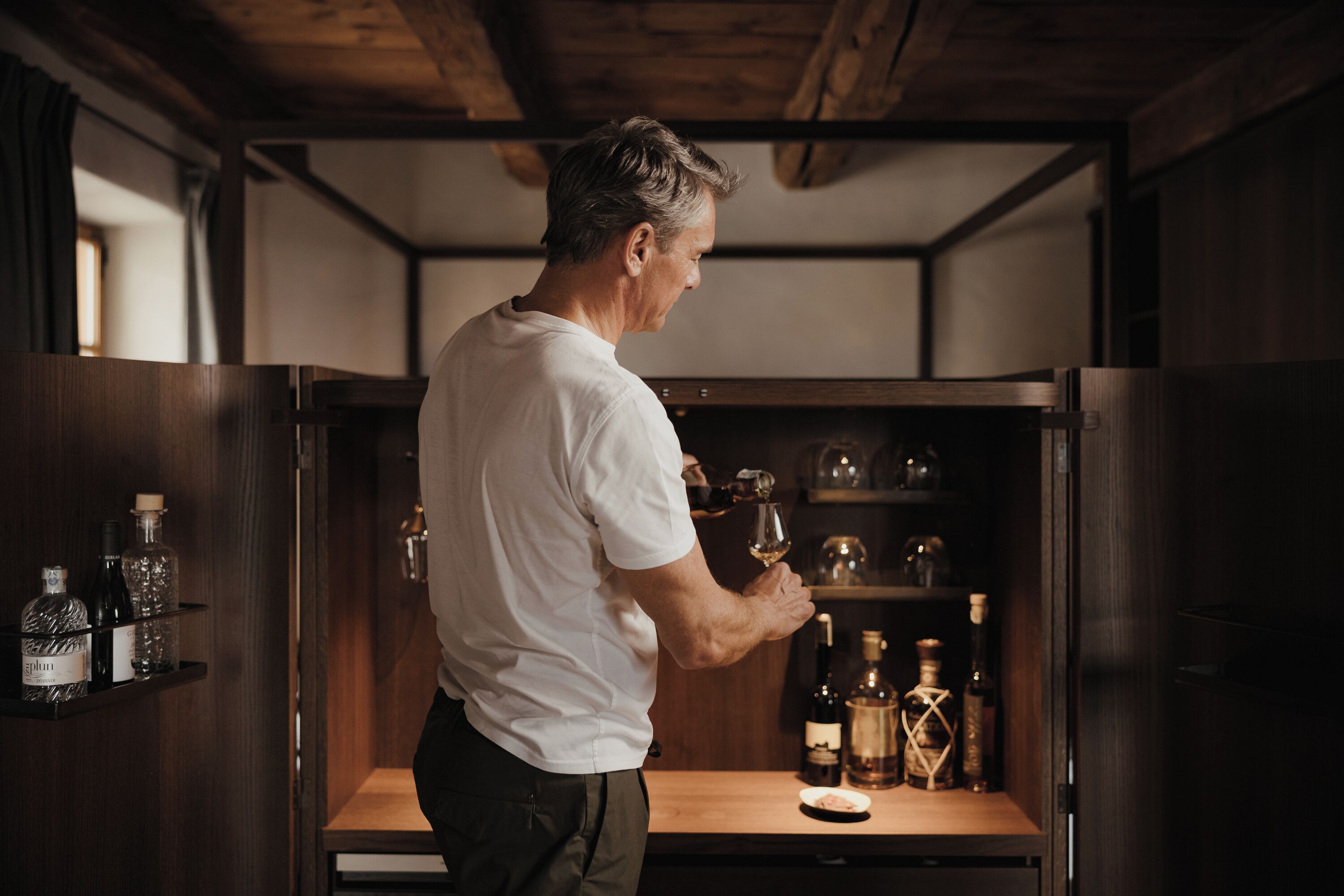 Un homme vêtu d’une chemise blanche verse du vin d’une bouteille dans un verre. On peut voir derrière lui des étagères remplies de différentes bouteilles et de verres.