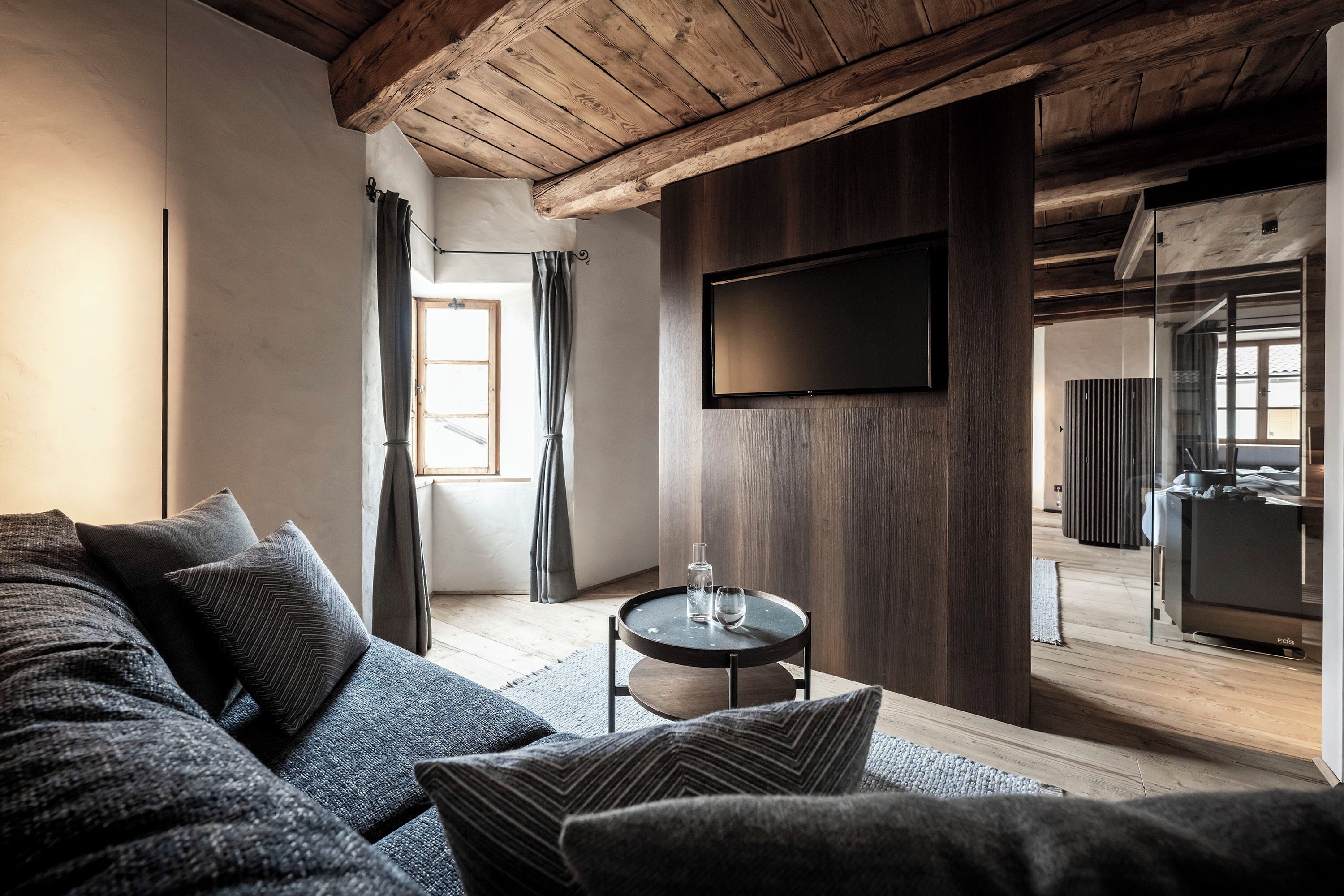 Een moderne woonkamer met een houten plafond, een flatscreen-televisie aan de wand, een donkerblauwe bank met gekleurde kussens en een klein rond tafeltje met een glazen blad.