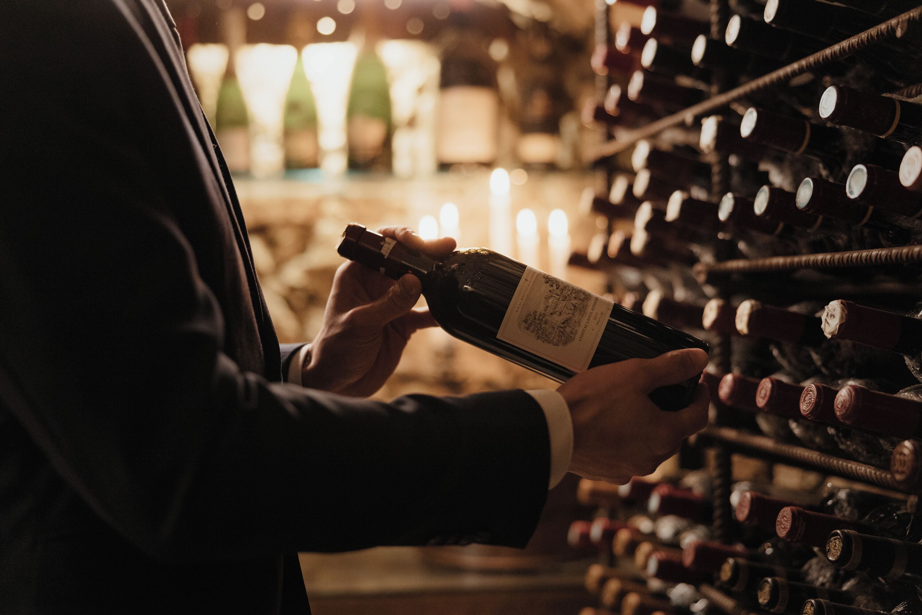 Una persona sostiene una botella de vino en una bodega, con hileras de botellas de vino en estanterías.