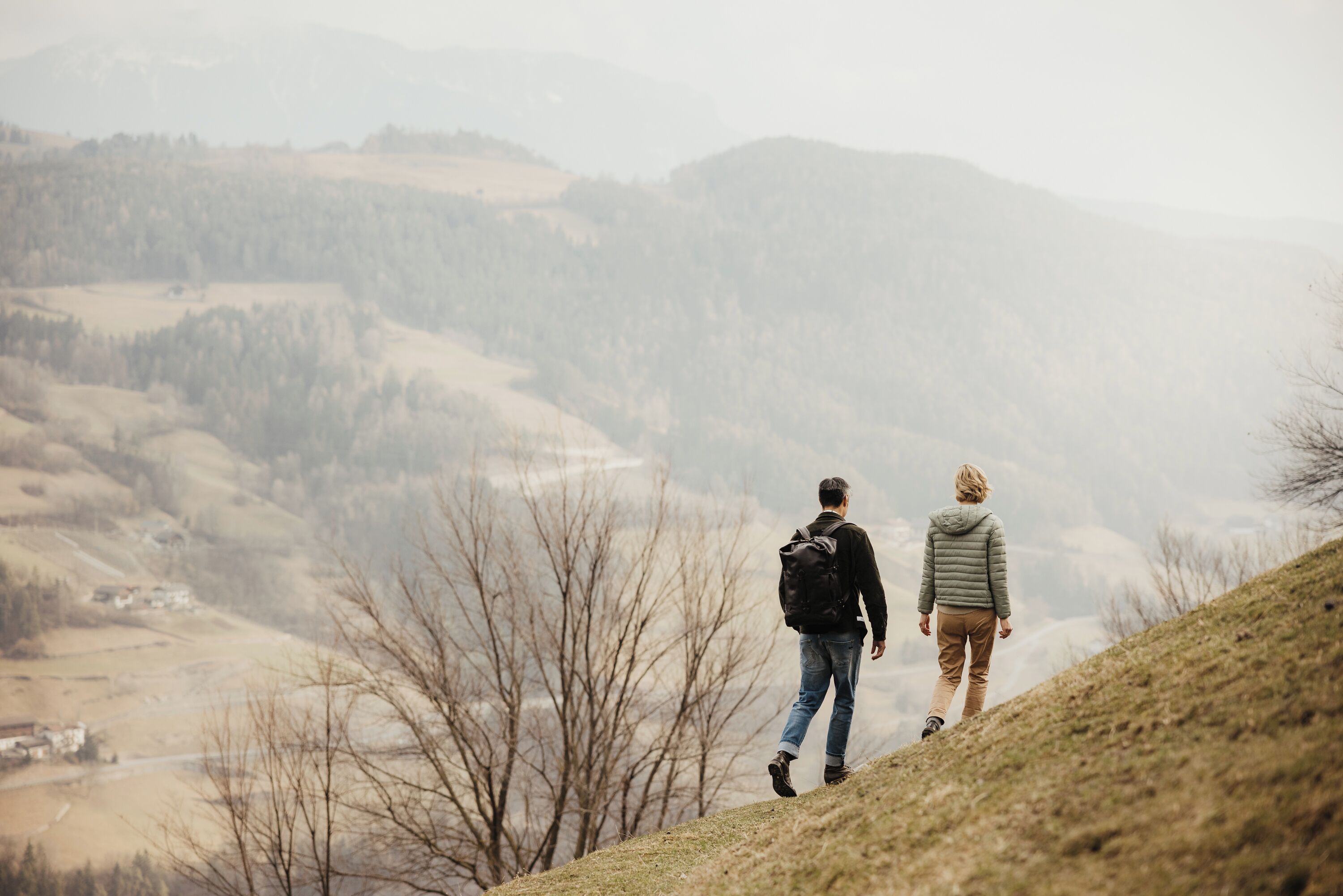 Två personer promenerar på en gräsklädd kulle med utsikt över en dal och berg i fjärran.