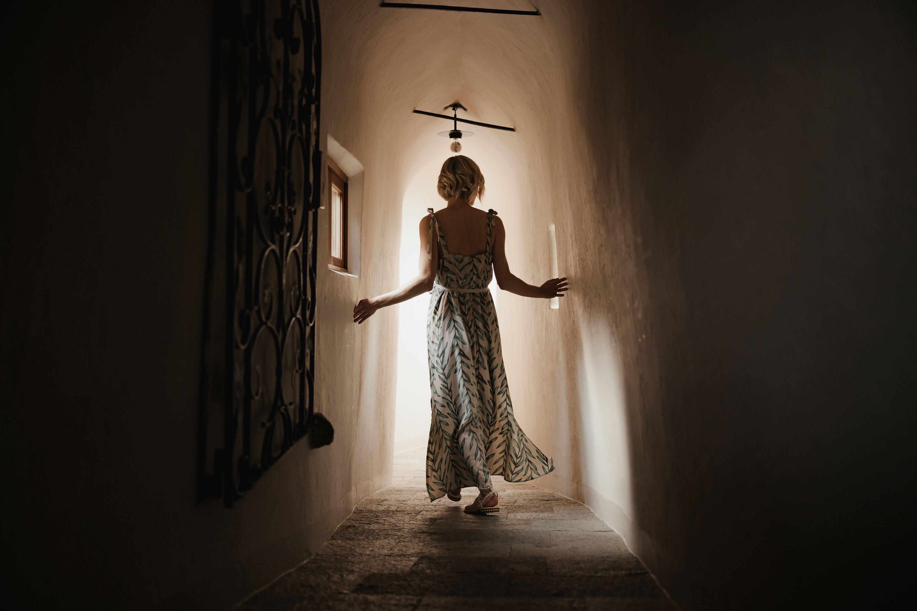 A person walking down a narrow hallway with a decorative iron railing on the left and a window on the right.