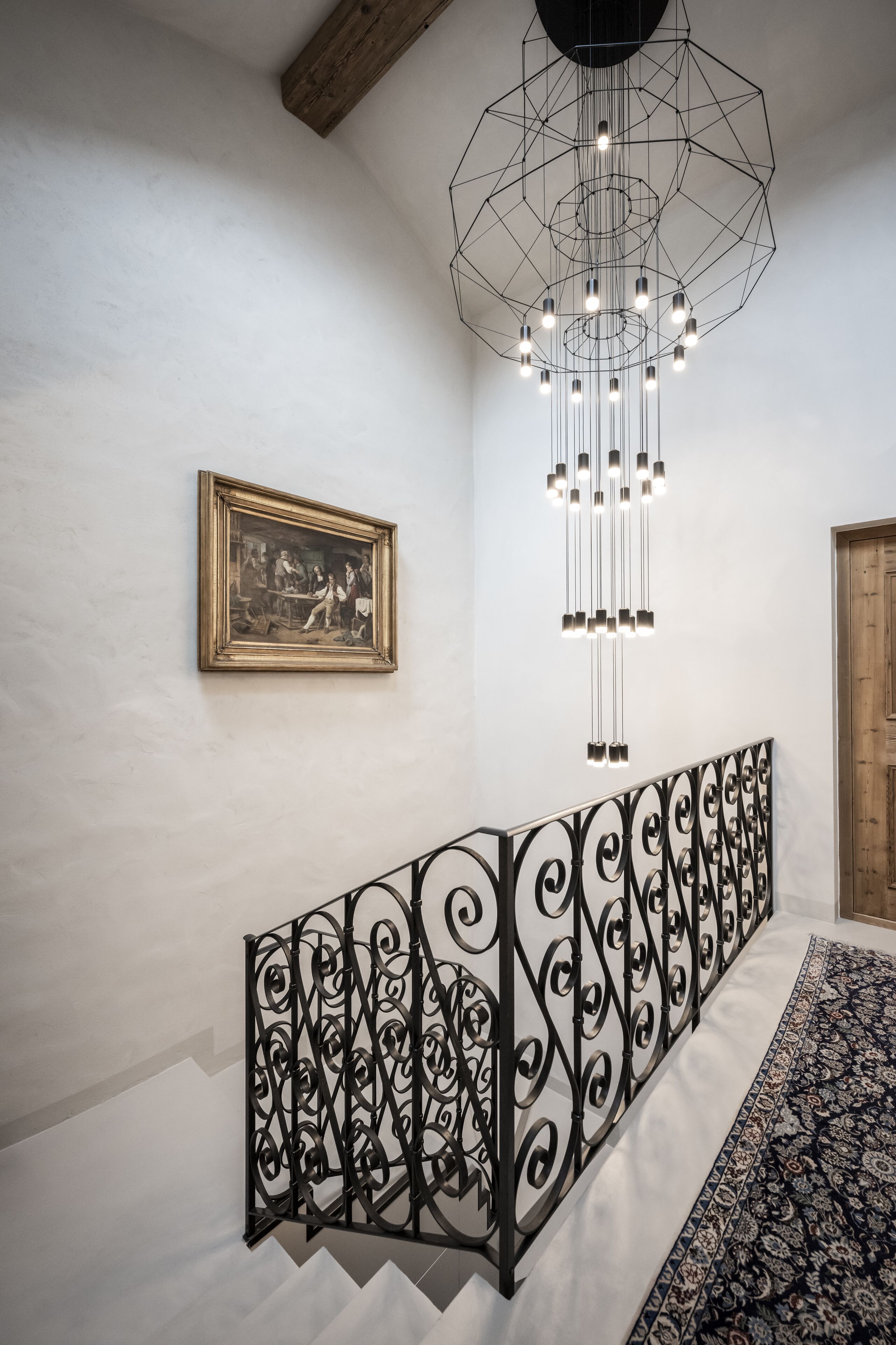 Una escalera con barandilla de hierro forjado negro, un candelabro geométrico y un cuadro en la pared.