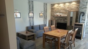 A modern living room with a sofa, a wooden dining table with chairs, a fireplace, and a stone wall. - Modern holiday house, relax in the countryside on one level (Karlshagen)