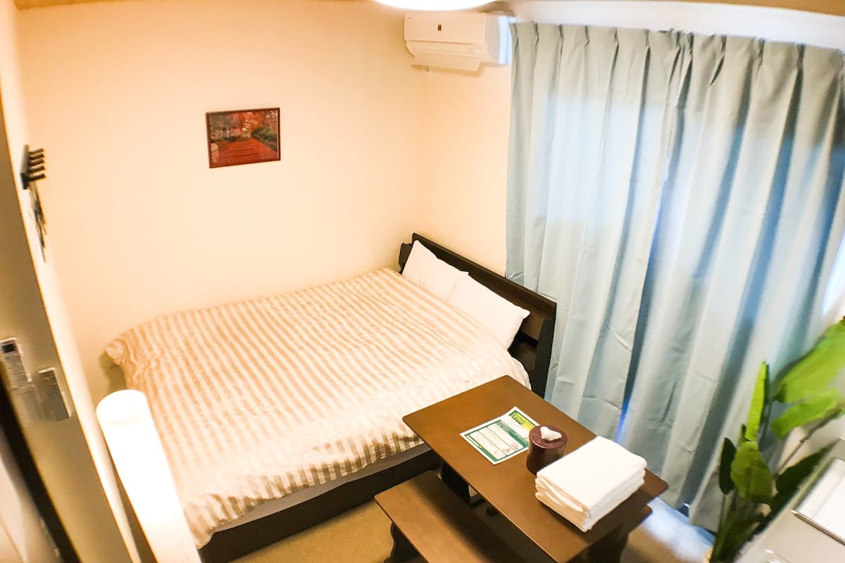 A single bed with a striped bedspread, a bedside table with a tissue box and a menu, a small bedside lamp, and a window with light blue curtains.