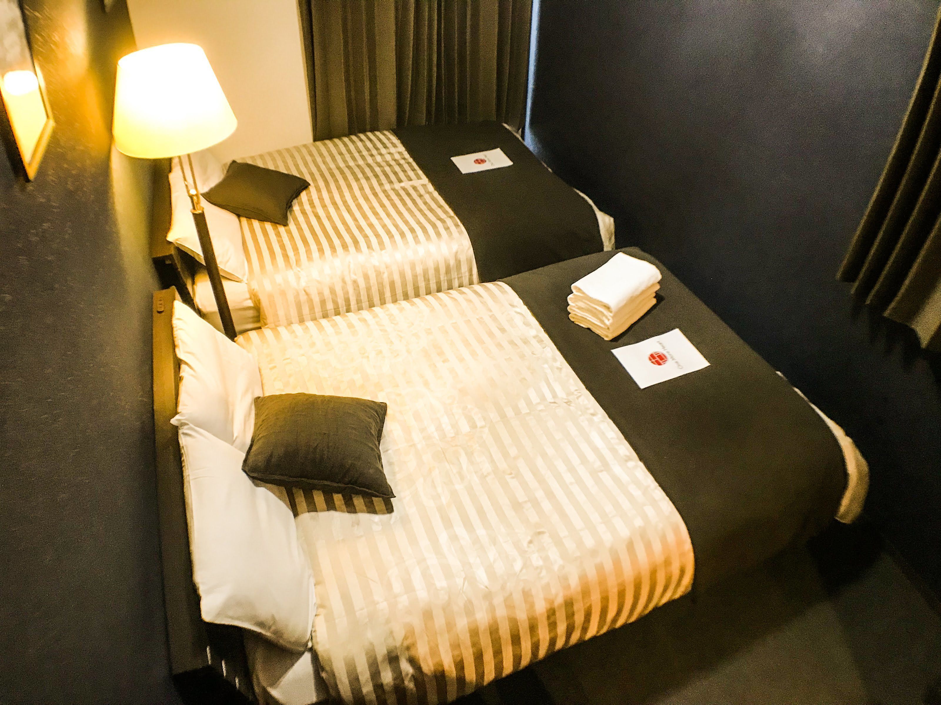 Une chambre d’hôtel avec deux lits, chacun doté de draps et d’oreillers à rayures. Une lampe se trouve sur le côté gauche de la pièce.
