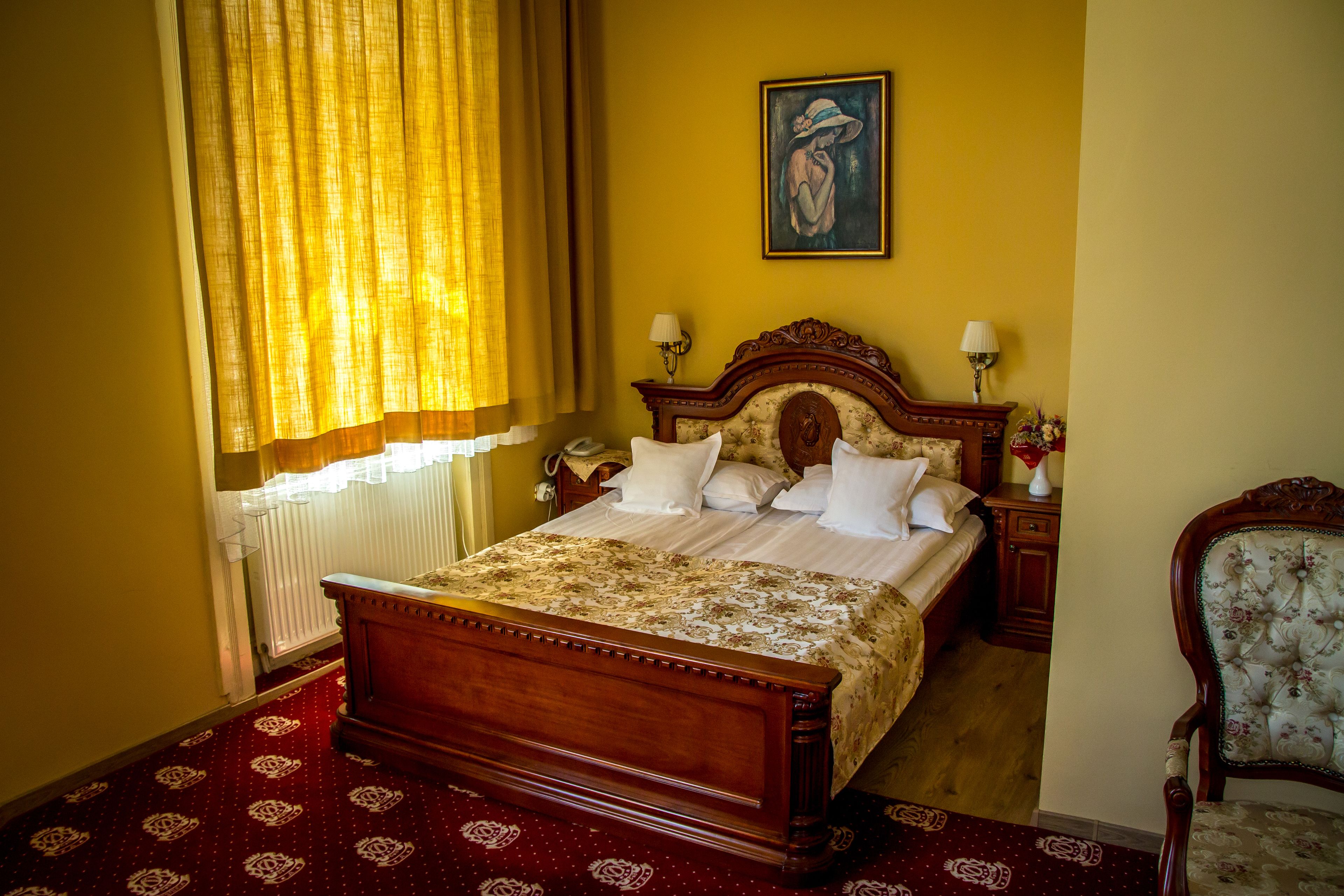 A bedroom with a wooden bed, two bedside tables with lamps, a chair, a painting on the wall, and a window with curtains.