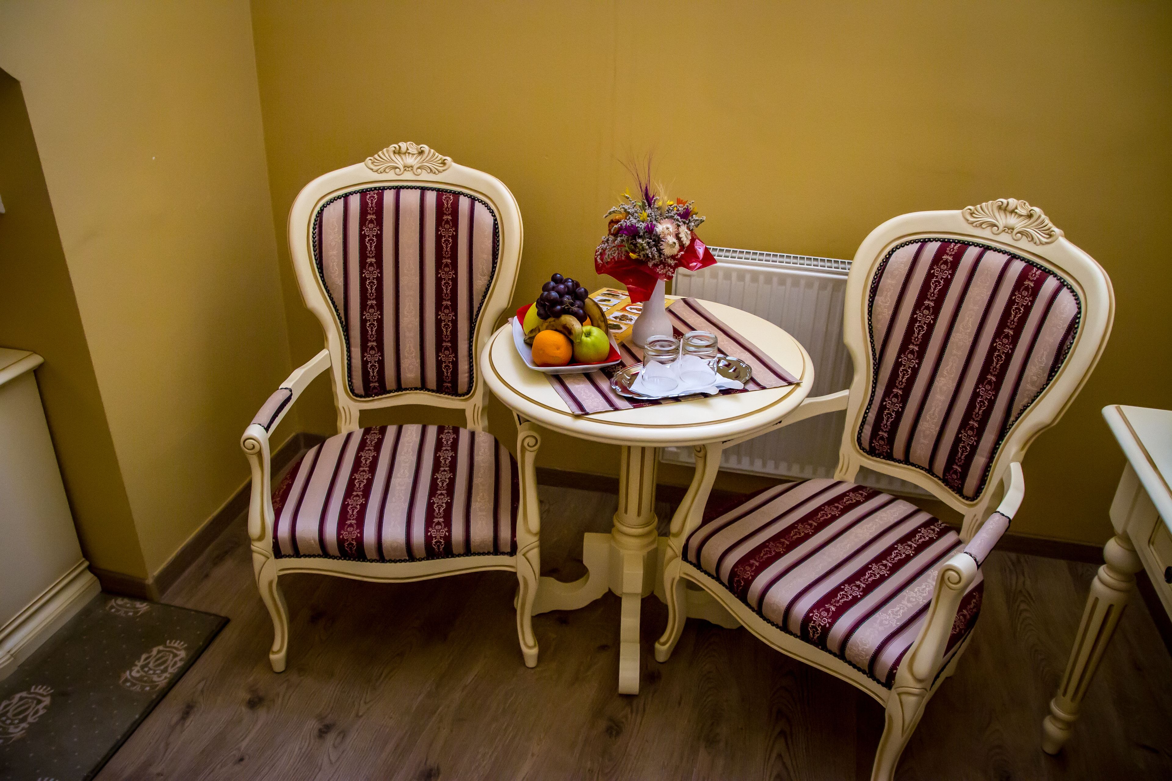 Two ornate chairs with striped upholstery, a small round table with a fruit plate, a vase with flowers, and a radiator in the background.