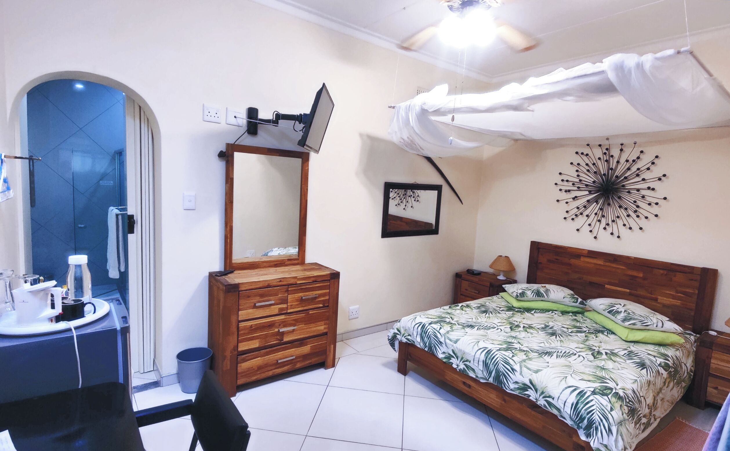 Ein Schlafzimmer mit einem hölzernen Bett, einem Nachttisch, einem Spiegel, einer Wanduhr und einem Deckenventilator.