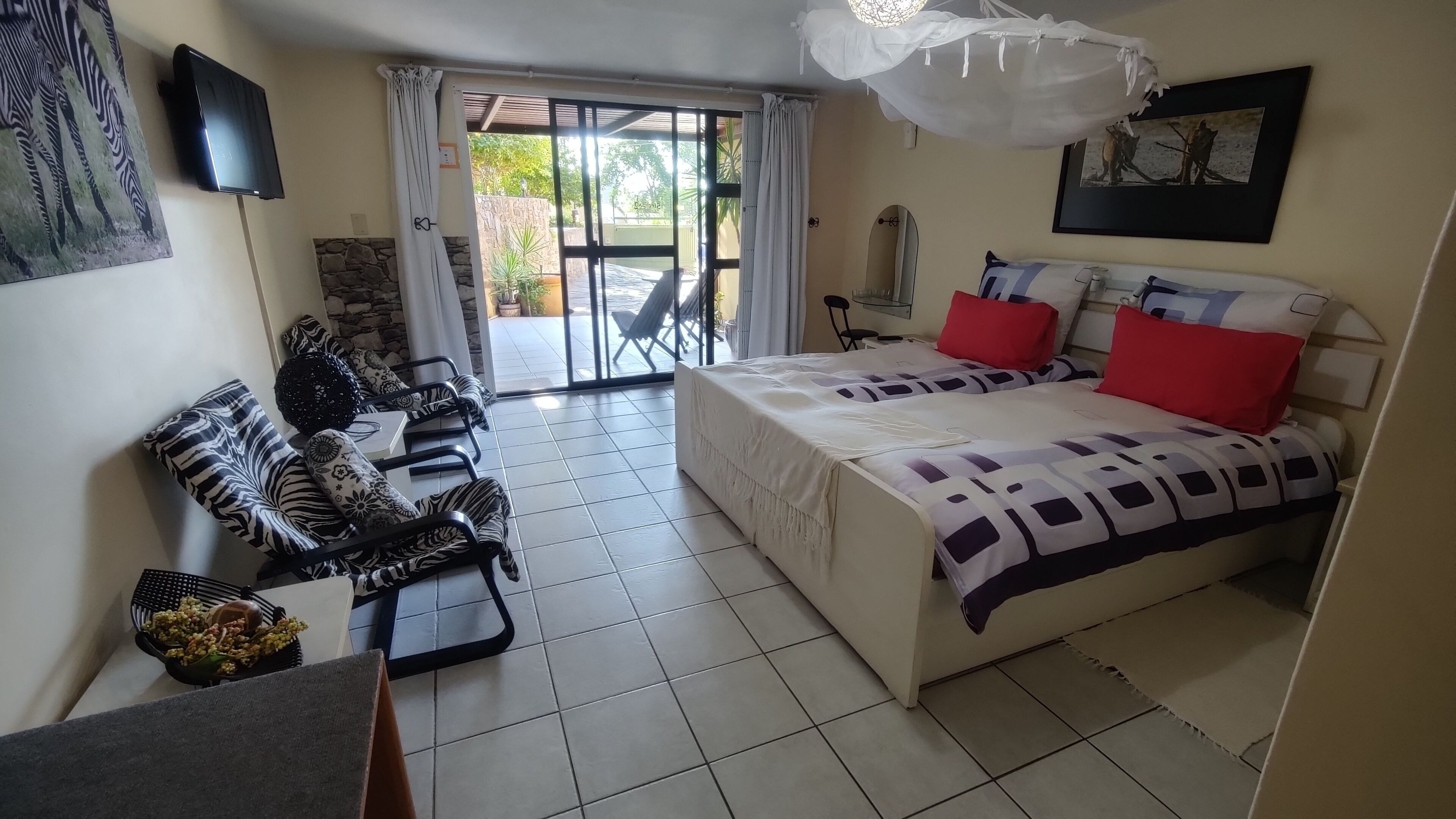 Habitación de hotel con una cama, dos sillas, una mesita pequeña, un ventilador de techo blanco y vistas al exterior a través de un vidrio.