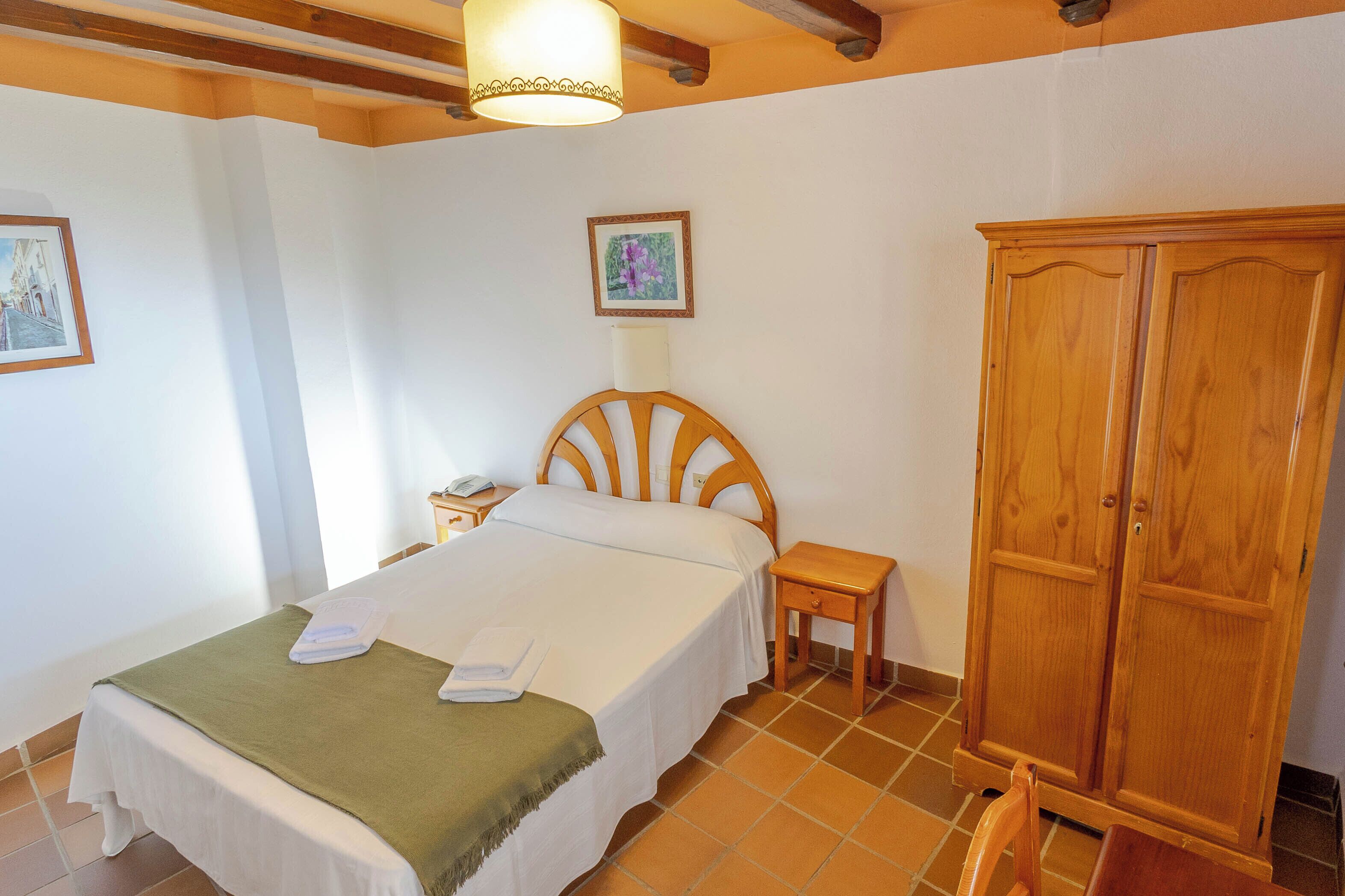 Une chambre à coucher comprenant un lit, une armoire en bois, une table de chevet et une lampe fixée au mur.