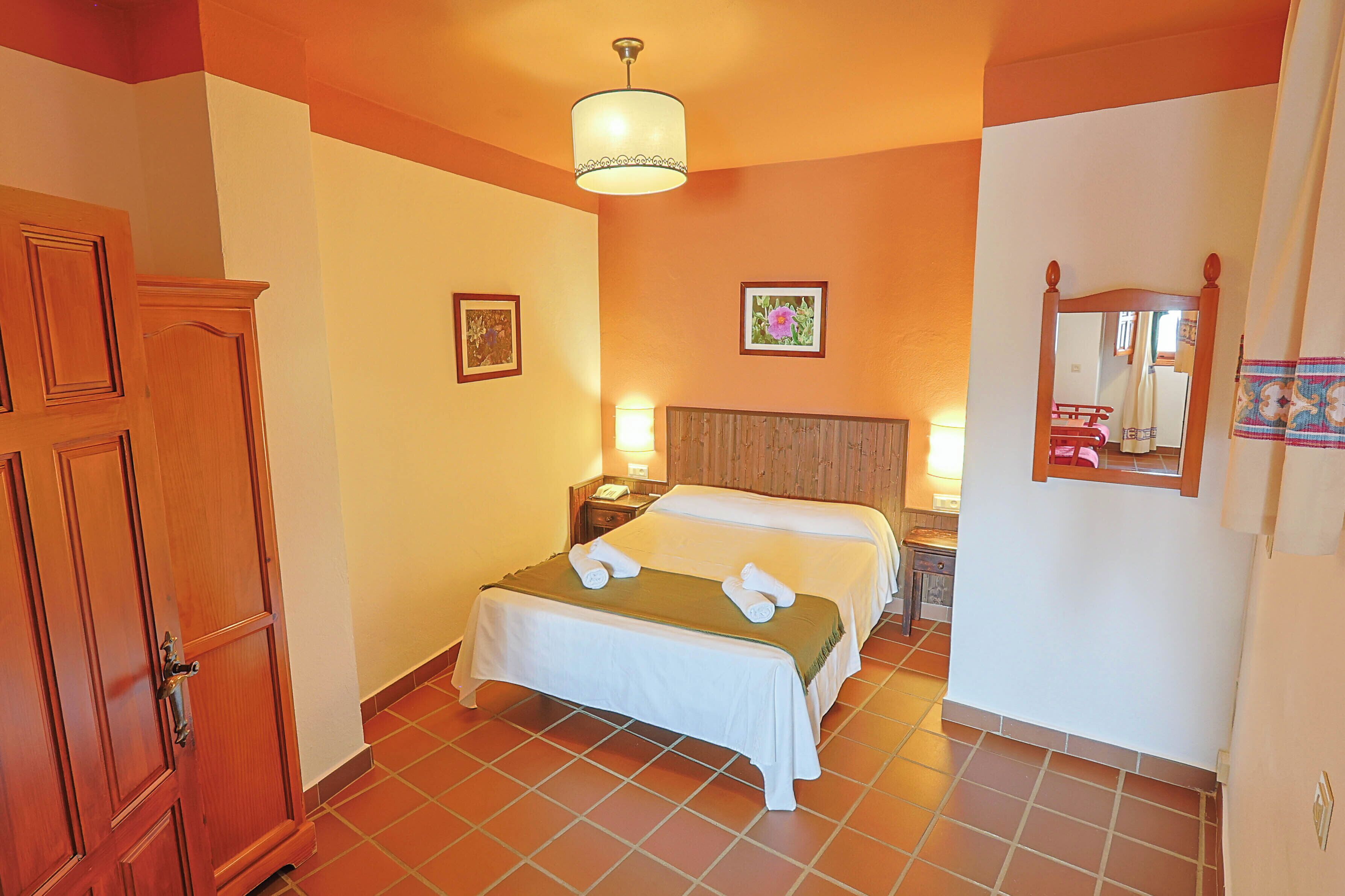 Una habitación de hotel con cama, armario de madera, mesita de noche con lámpara y espejo en la pared.