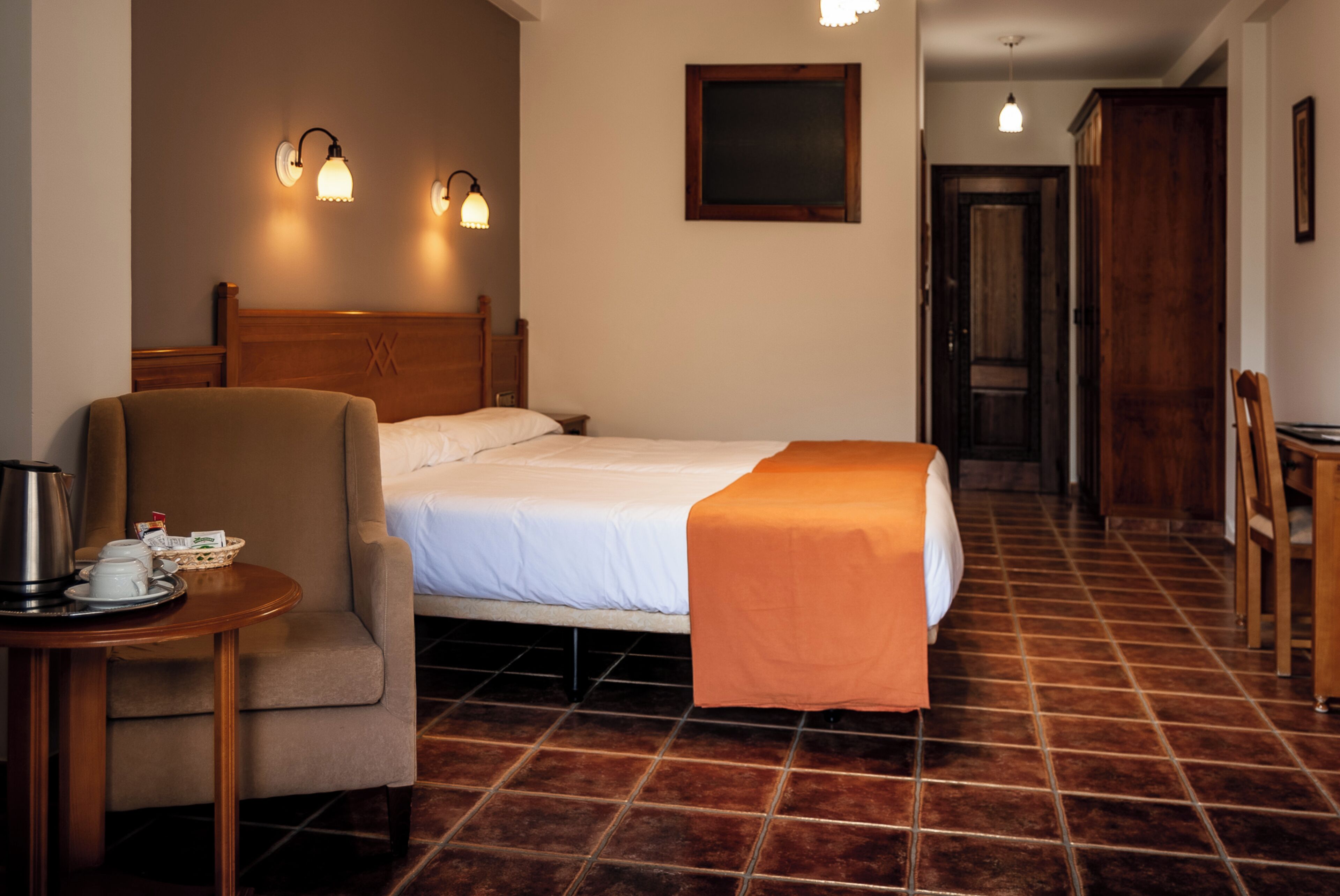 A hotel room with a bed, a chair, a table with a kettle and cups, and a wooden headboard.