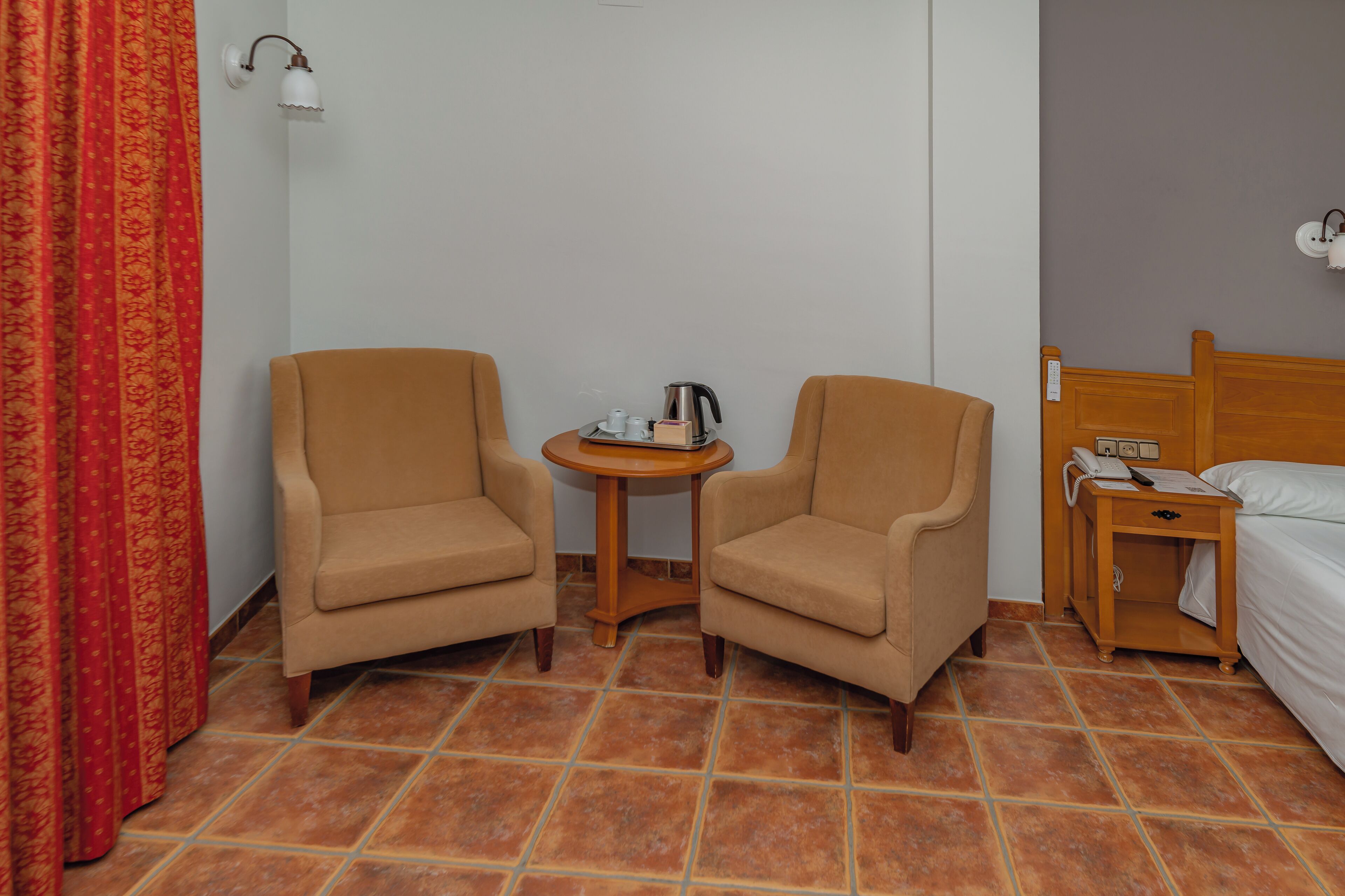A hotel room with two armchairs, a small round table with a kettle, a bedside table with a phone, and a bed.