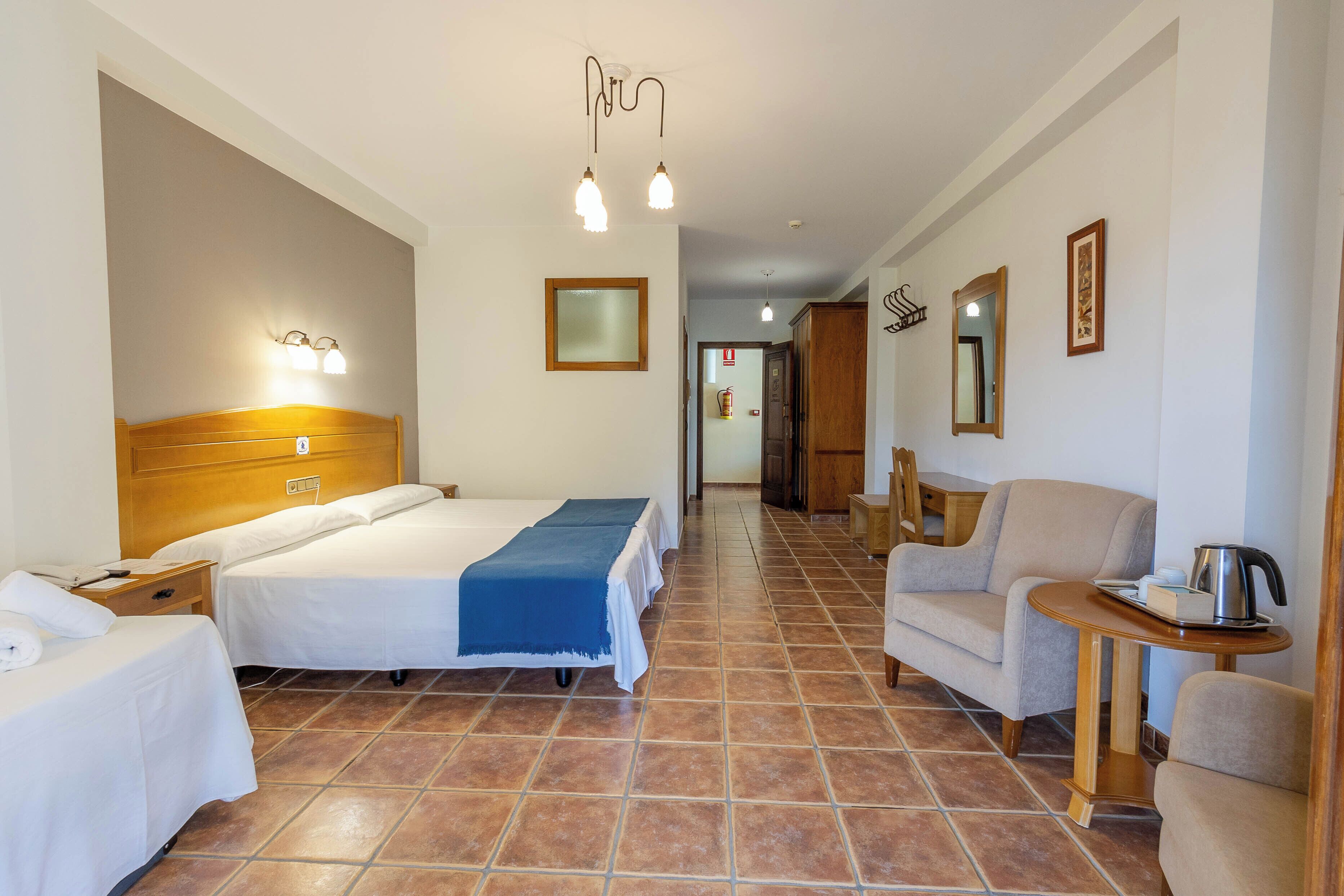 Habitación de hotel con dos camas, un sofá, una mesita con hervidor y un espejo en la pared.
