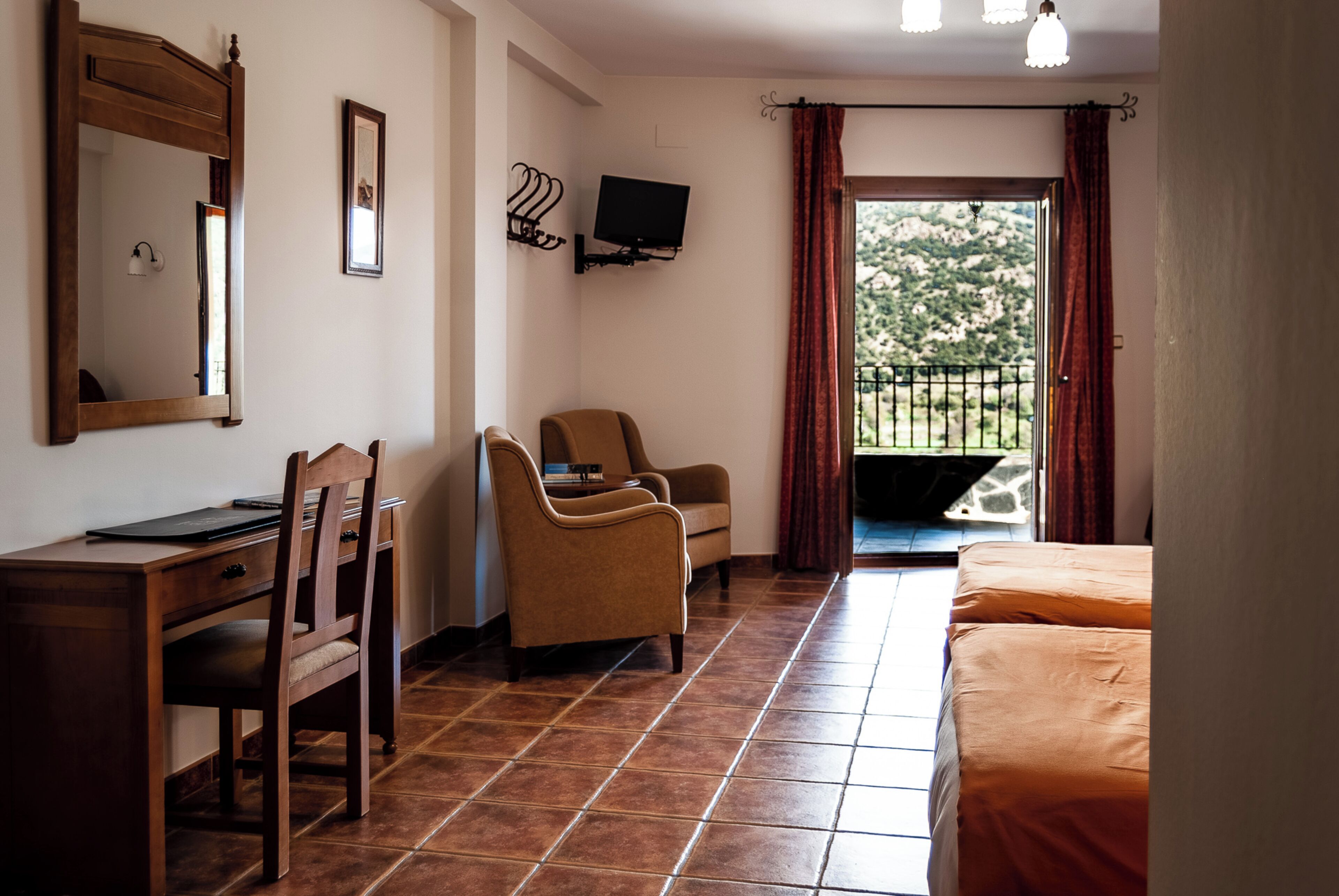 Habitación de hotel con cama, escritorio con silla, televisor montado en la pared y balcón con vistas al verdor.