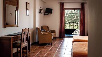 Habitación de hotel con cama, escritorio con silla, televisor montado en la pared y balcón con vistas al verdor.