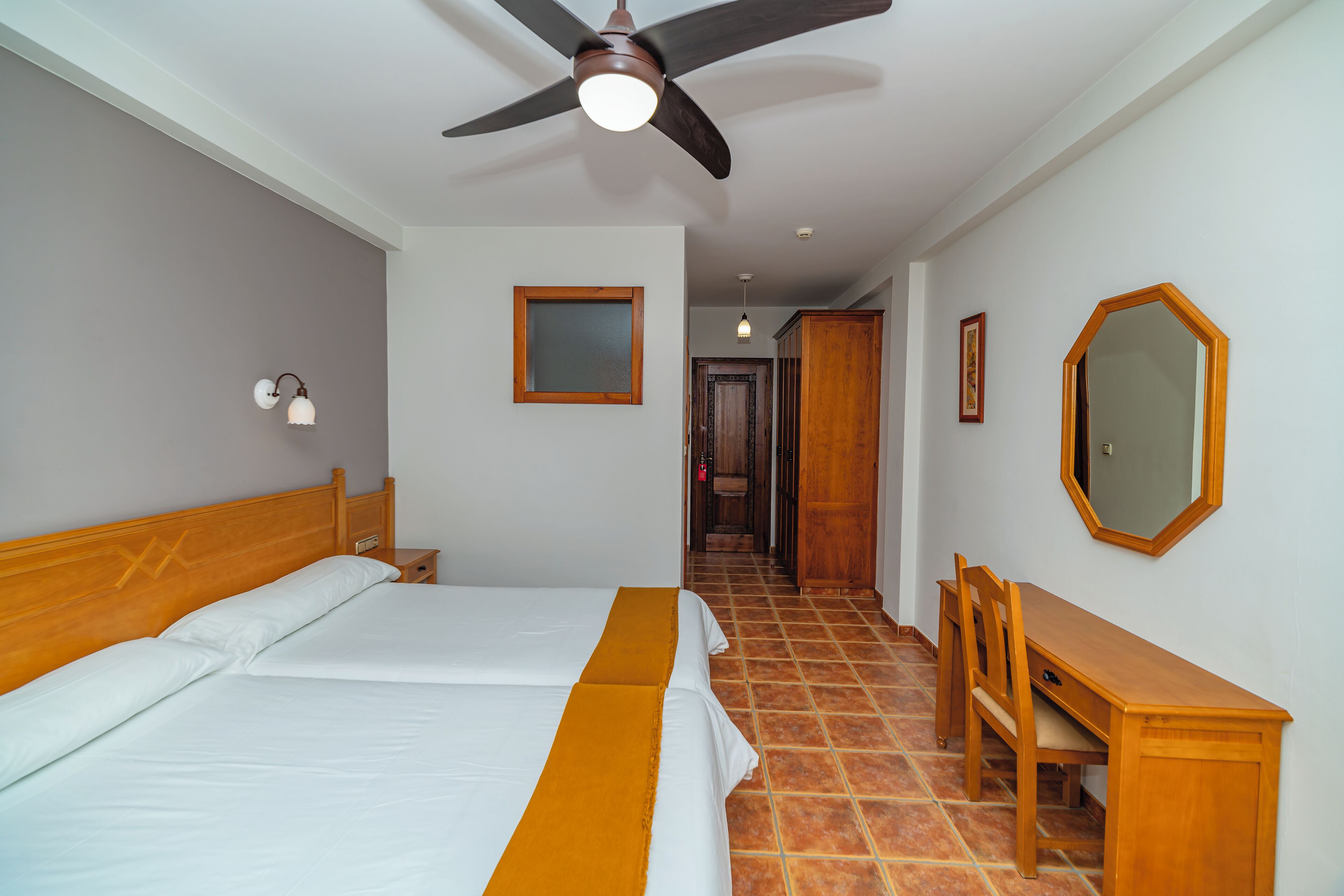 Une chambre d’hôtel équipée d’un lit, d’un bureau, d’une chaise, d’un ventilateur de plafond et d’un miroir au mur.