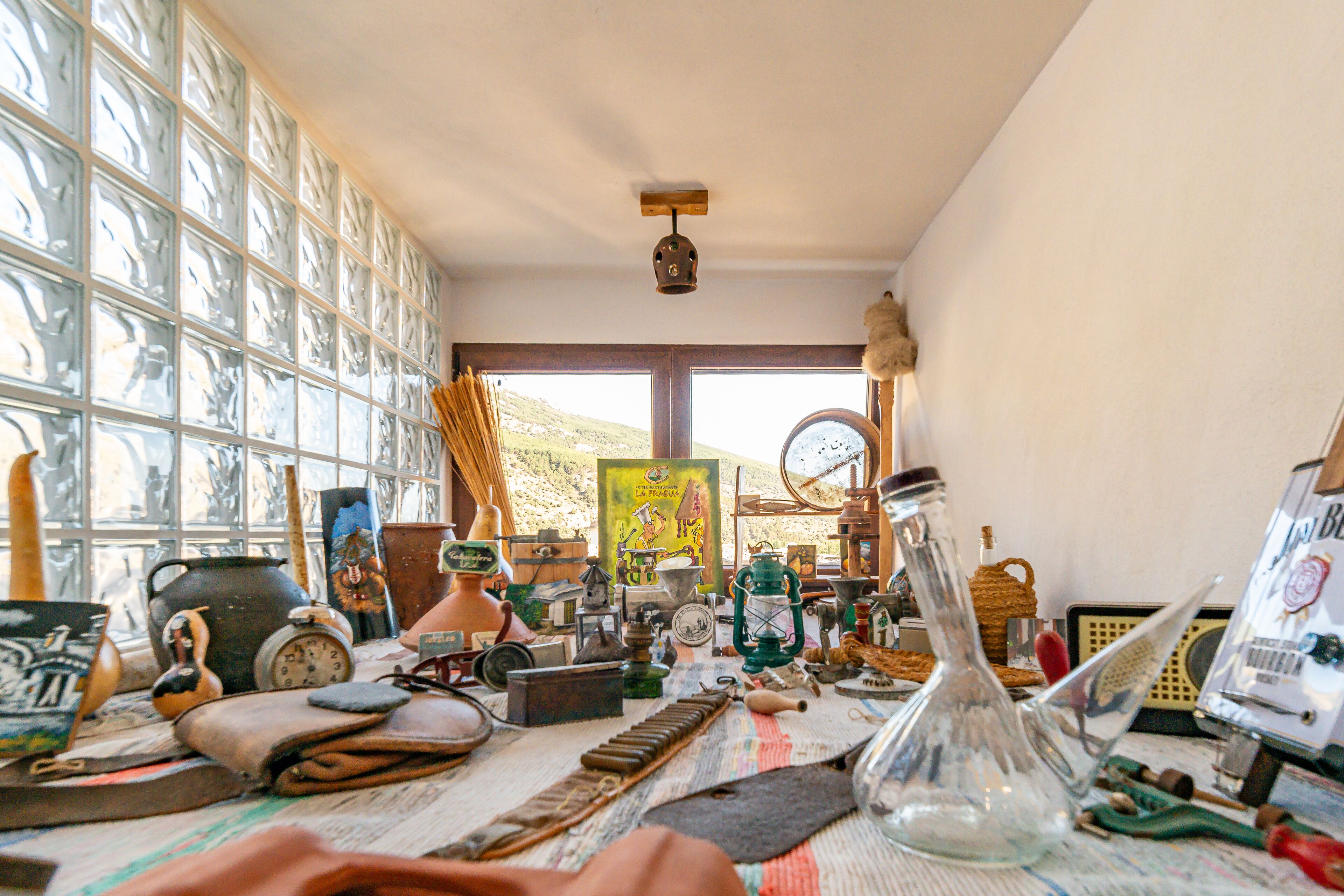 Una habitación con ventanas amplias, diversos objetos antiguos sobre una mesa y un cuadro en la pared.