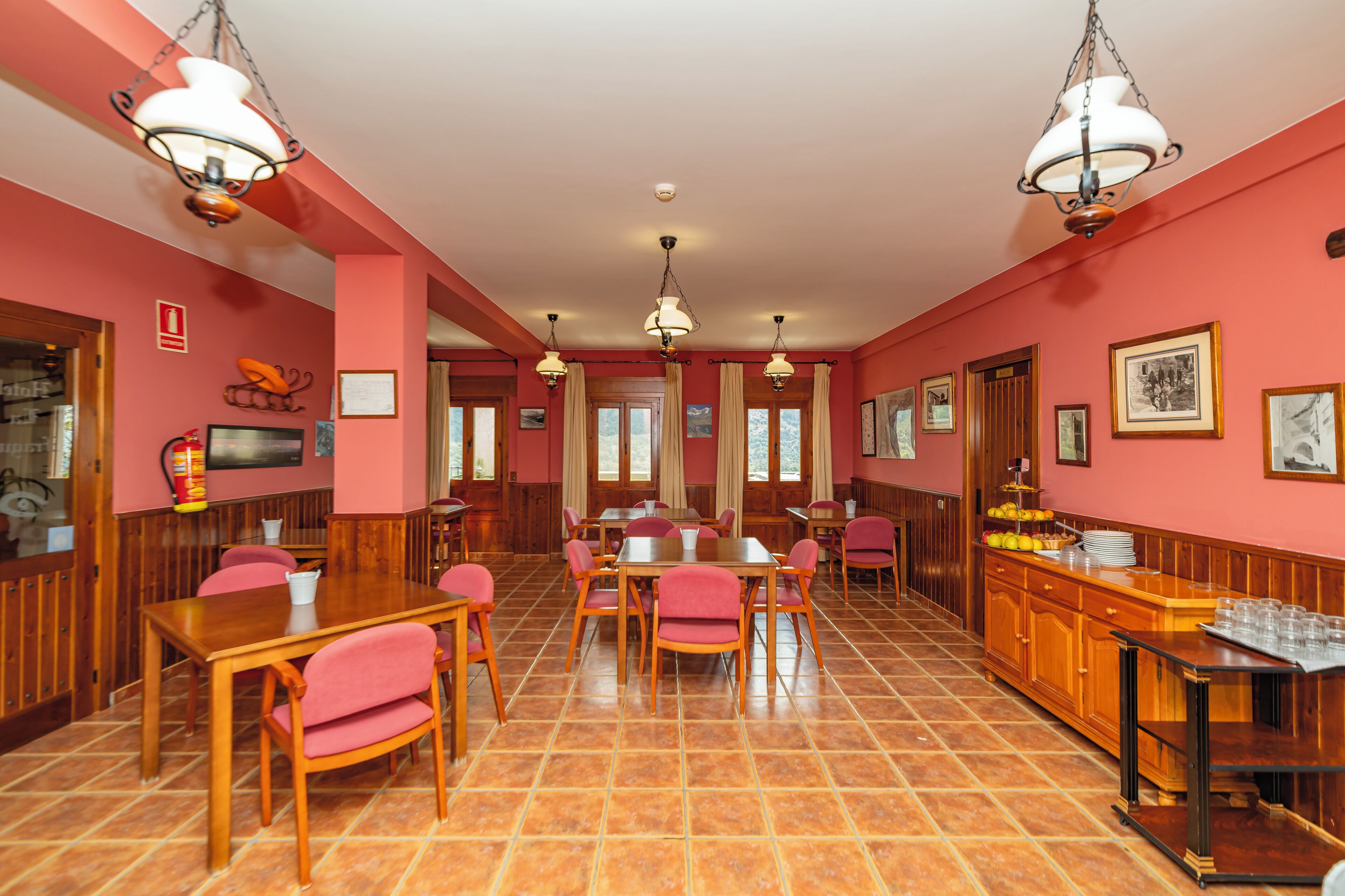 Un espace repas avec des tables et des chaises en bois, un buffet garni de plats et de boissons, et des lampes suspendues.