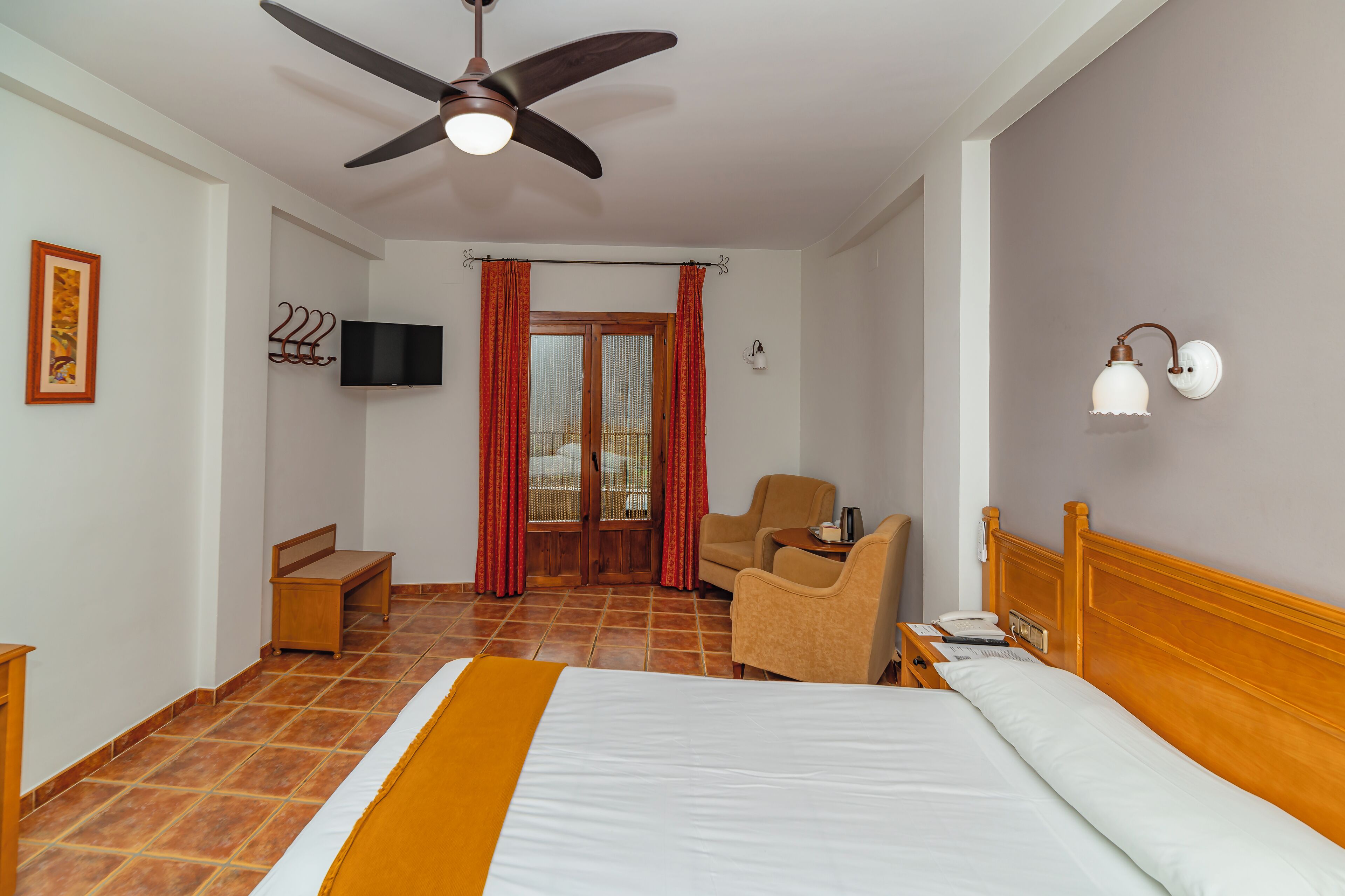 Habitación de hotel con cama, una silla, escritorio, televisor, ventilador de techo y ventana con cortinas.