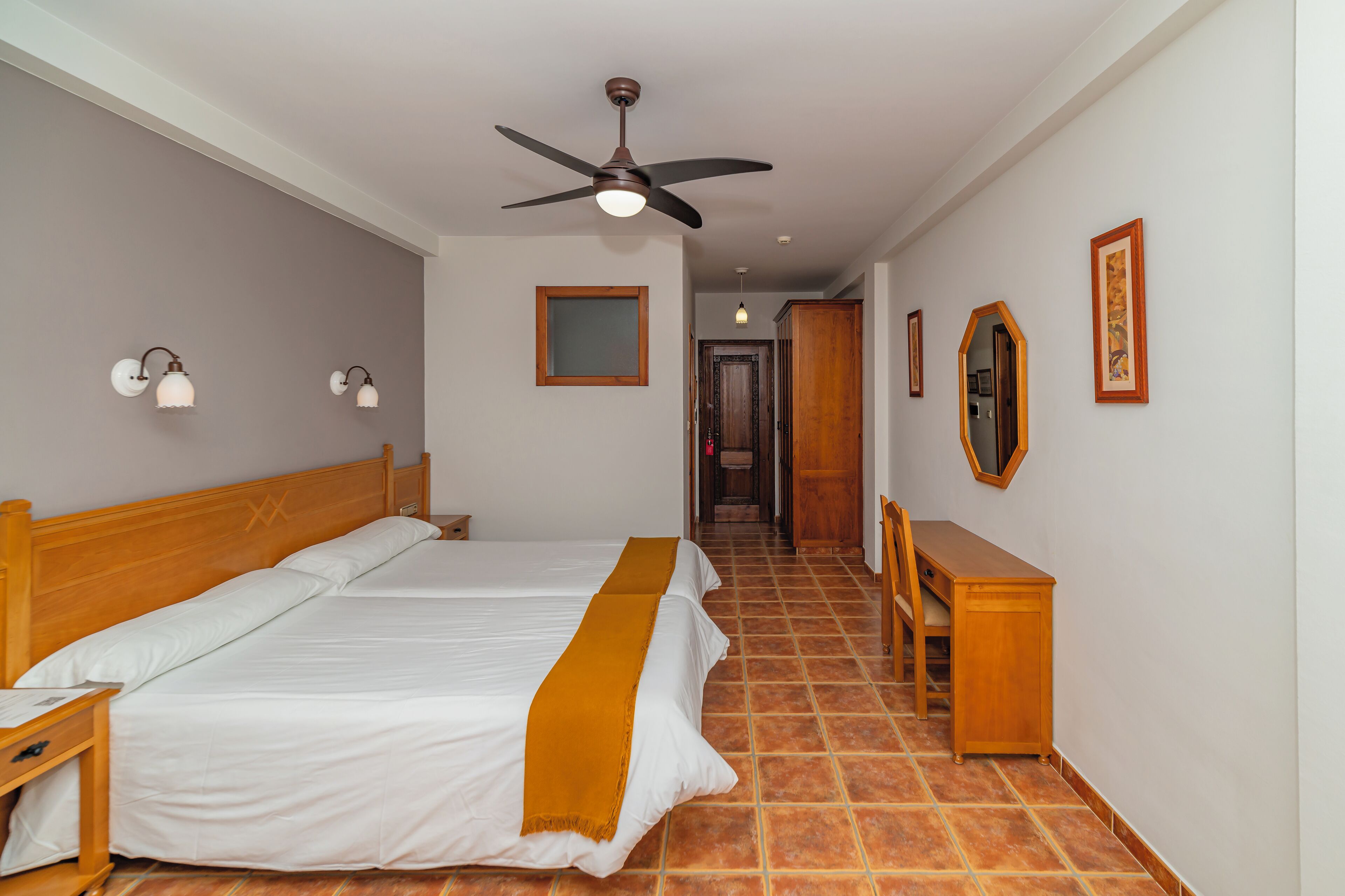 Una habitación de hotel con una cama grande, una mesita de noche de madera, un escritorio y un ventilador de techo.