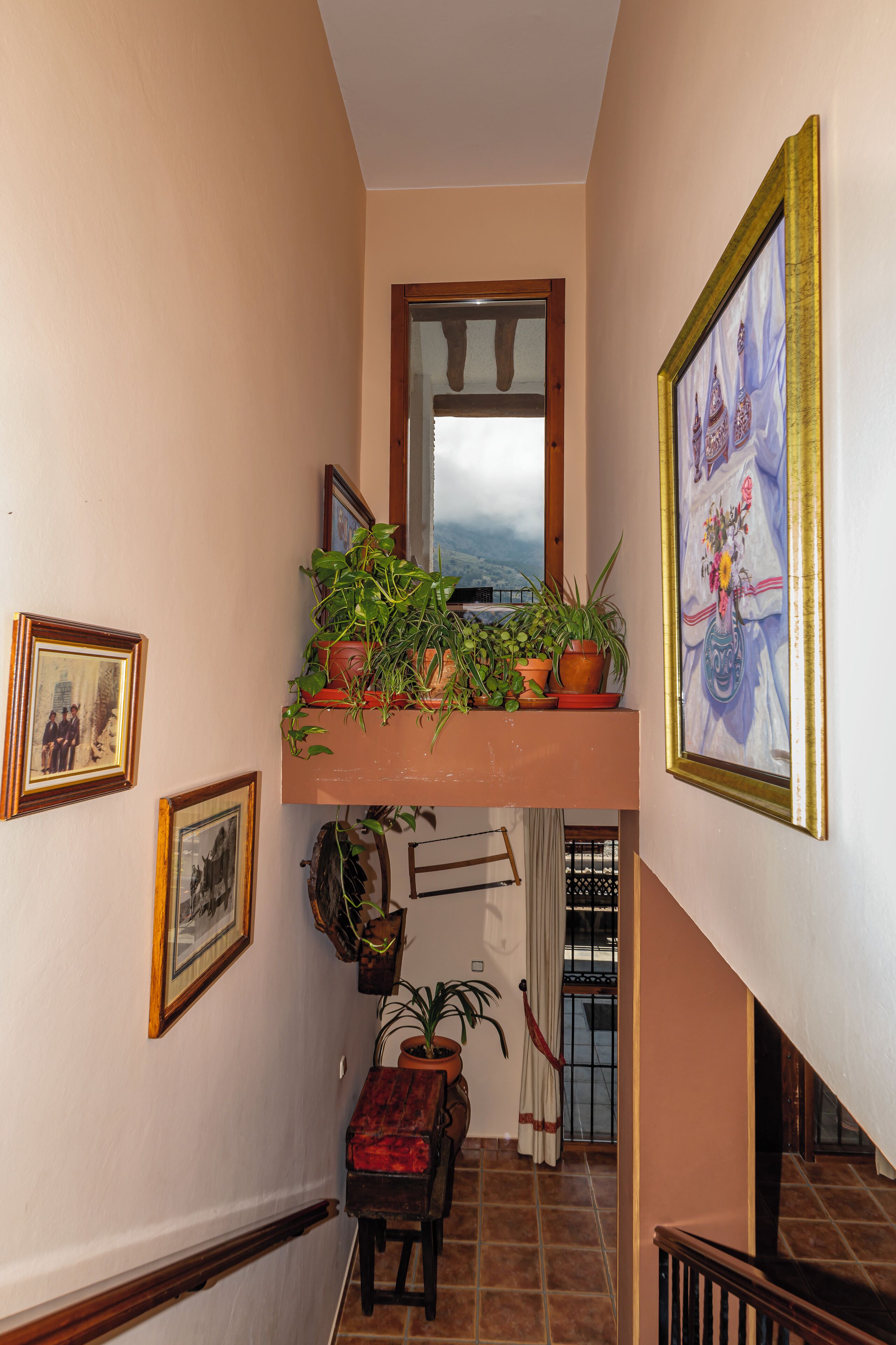A narrow hallway with a wooden staircase, a balcony with potted plants, and framed pictures on the walls.