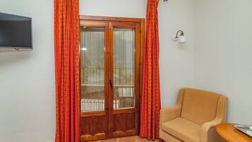 Una habitación con puerta de madera, cortinas rojas, un sofá color beige, una mesita y un televisor fijado a la pared.