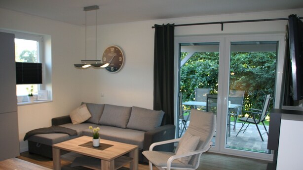 Una sala de estar moderna con un sofá, una mesa de centro y una silla. Hay un reloj en la pared y una ventana con cortinas.