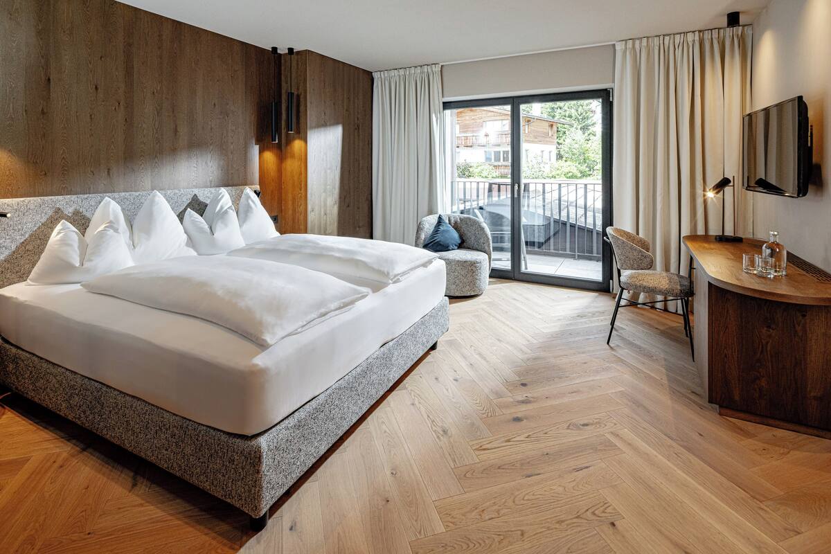 A modern hotel room with a large bed, a desk, a chair, and a balcony with a view.