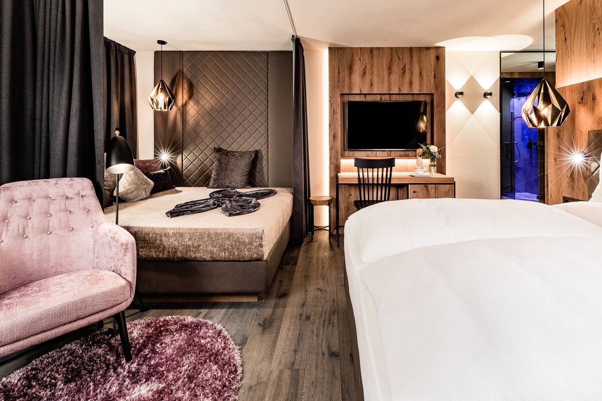 A modern hotel room with a large bed, a desk with a chair, a pink armchair, and a large mirror.