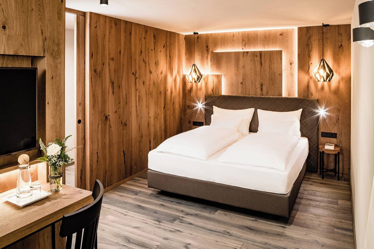 A modern bedroom with a wooden wall panel, a bed with white linens, a desk with a vase, and two wall-mounted lights.