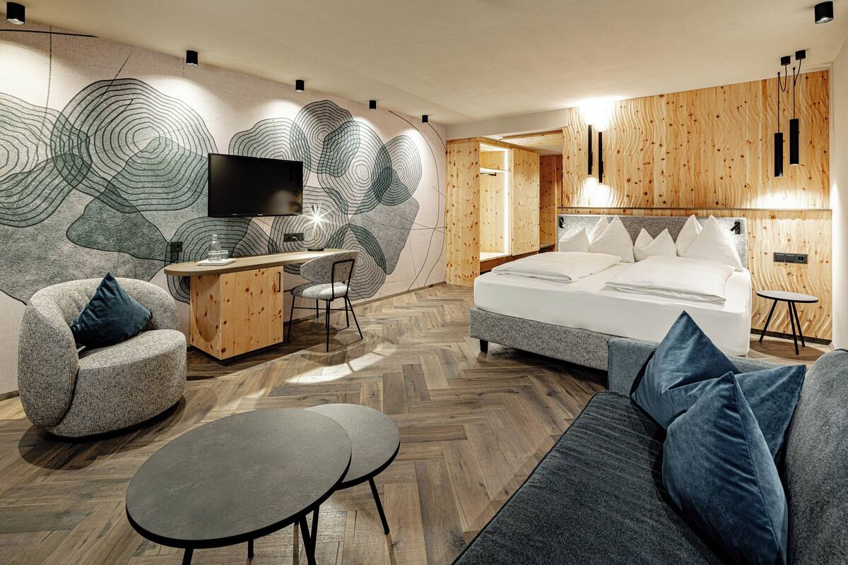 A modern hotel room with a large bed, a desk with a TV, a sofa, and a herringbone wood floor.