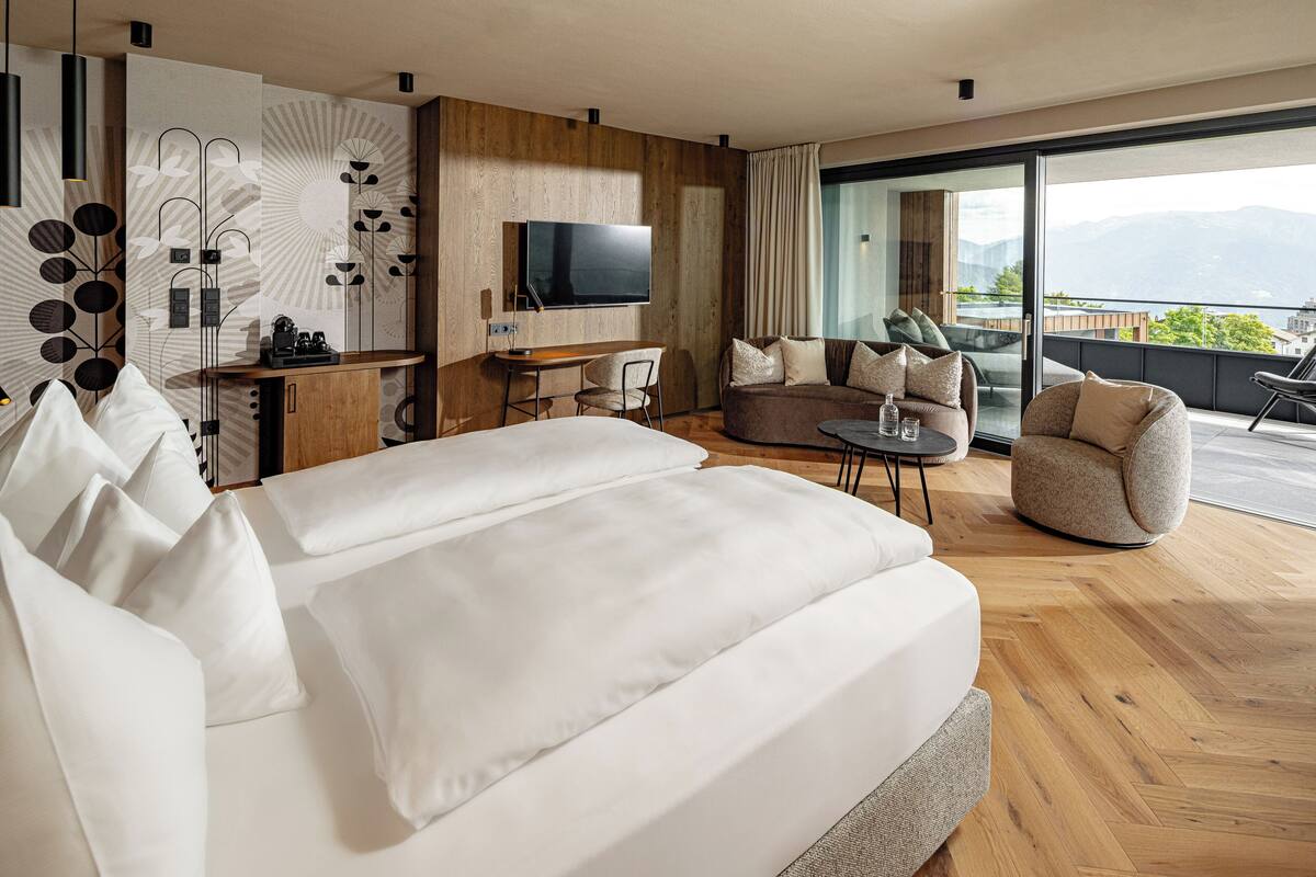 A modern hotel room with a large bed, a desk with a flat-screen TV, a sofa, and a balcony with a mountain view.