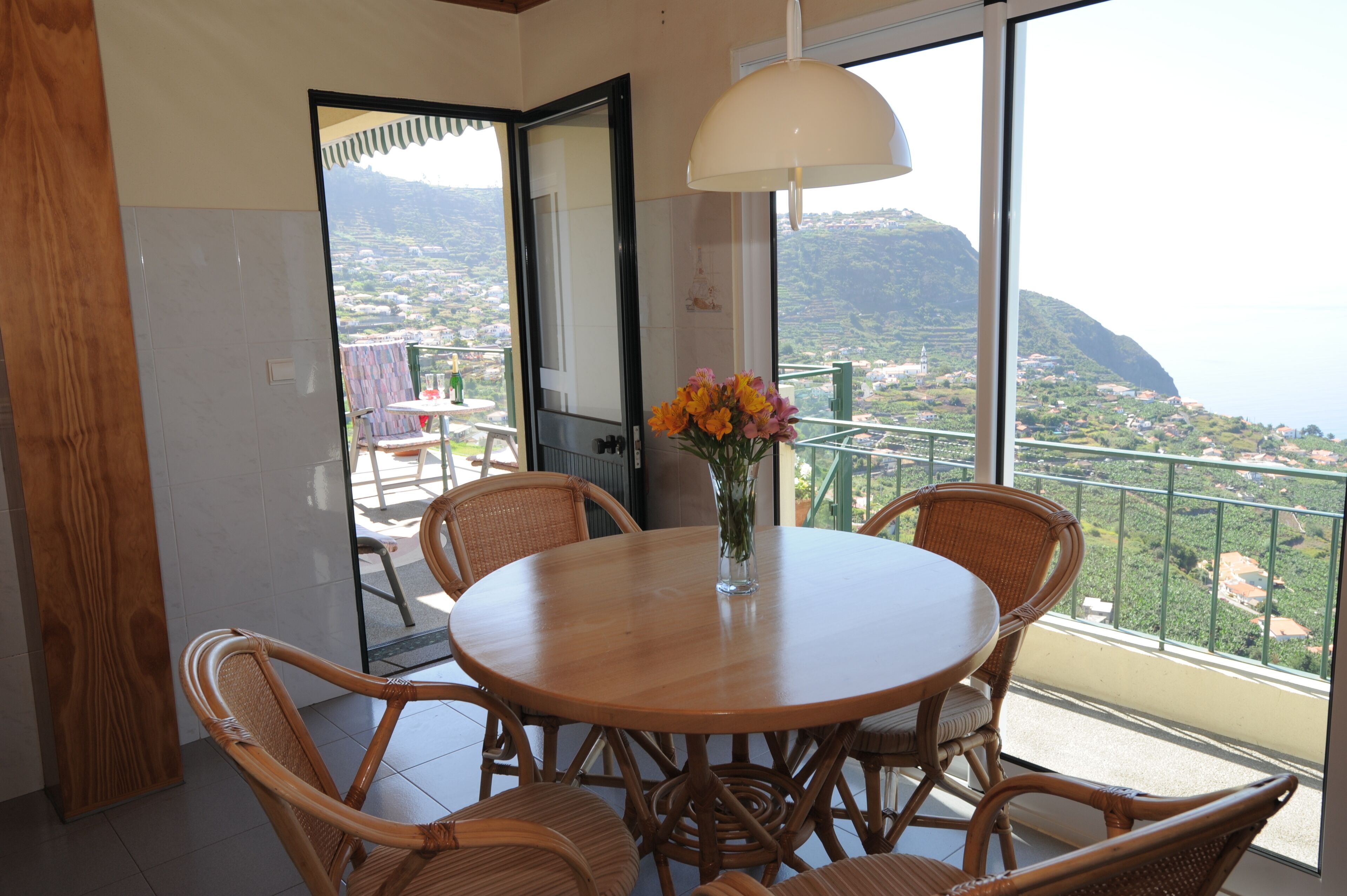 Un balcón con sillas de mimbre y una mesa redonda, un jarrón con flores y vistas al paisaje costero.
