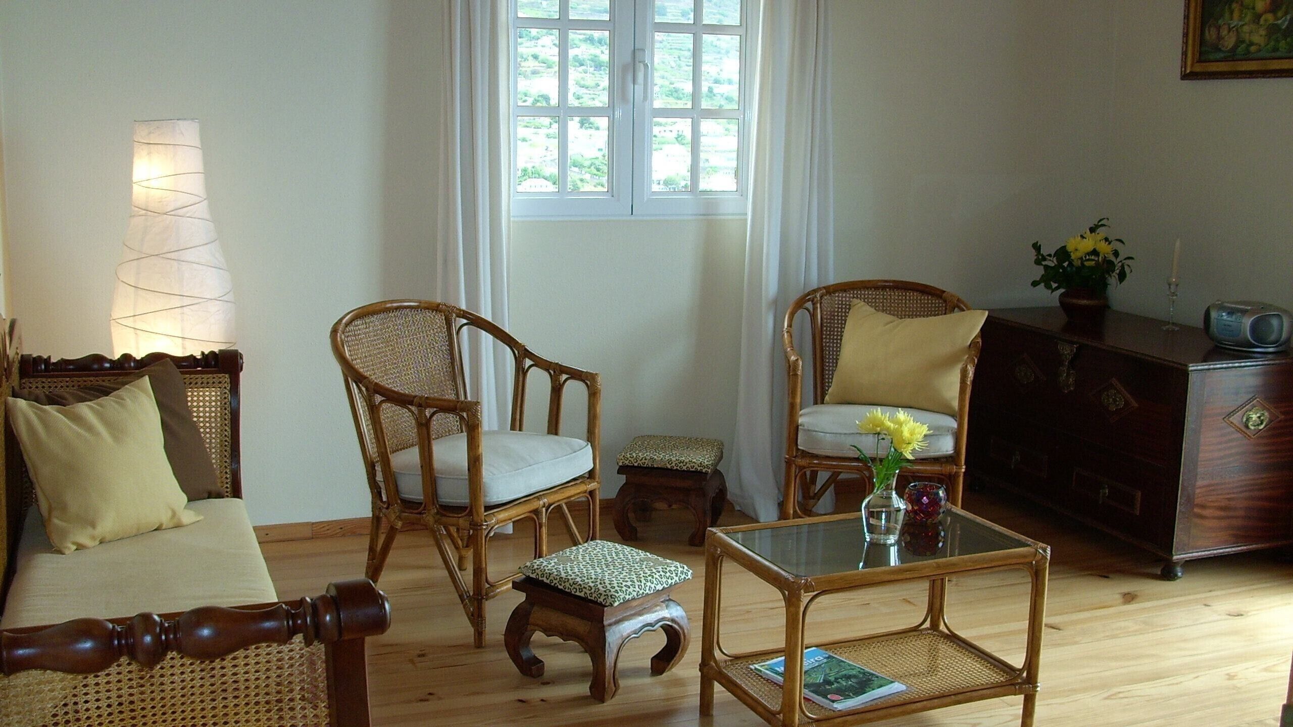 A room with a sofa, two wicker chairs, a small table with a vase of flowers, a wooden cabinet, and a window with a view of greenery.