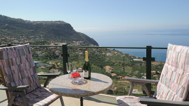 A balcony with two chairs, a table, and a bottle of champagne. The view includes a coastal landscape with houses and greenery. - 60 sqm apartment with 2 bedrooms and fantastic sea views (Arco da Calheta)