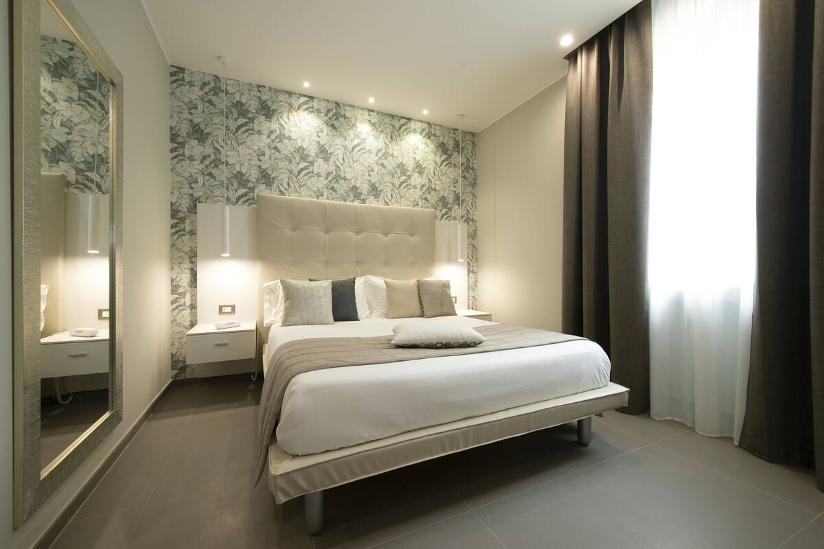 Un dormitorio moderno con una cama grande, una mesita de noche, un espejo y una pared cubierta de papel tapiz floral.