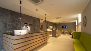 Una zona de recepción con un mostrador de madera, una computadora en el mostrador, un mostrador con un reloj, un mostrador con un televisor, un mostrador con una planta, un mostrador con una ventana, un mostrador con una silla, un mostrador con una mesa, un mostrador con un sofá, varios mostradores con plantas.