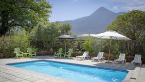 Una piscina con sillas de descanso y sombrillas, rodeada de vegetación y con una montaña al fondo.