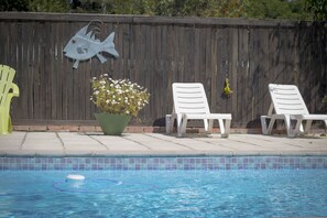 A swimming pool with a wooden fence, a fish decoration, and two white lounge chairs.