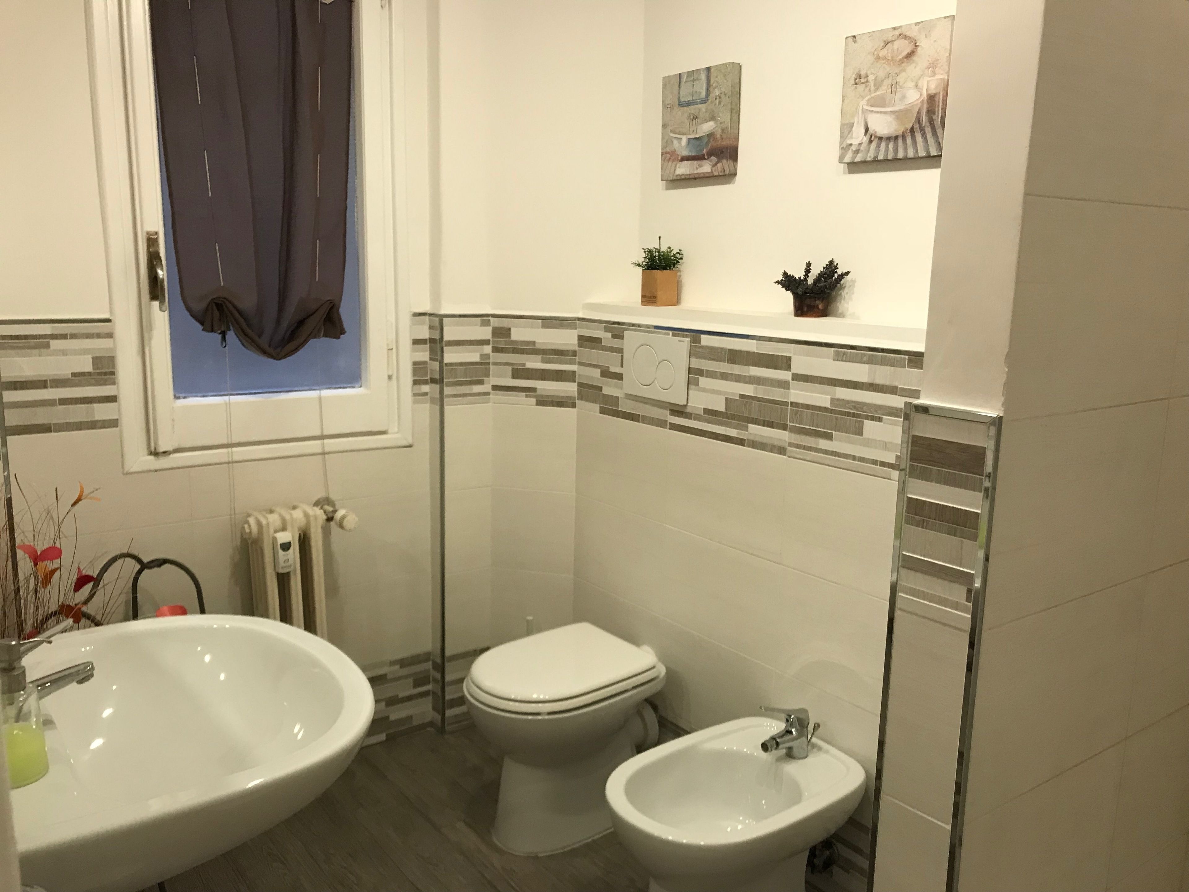 A bathroom with a double sink, toilet, and bidet. There is a window with a curtain, a radiator, and two wall-mounted paintings.