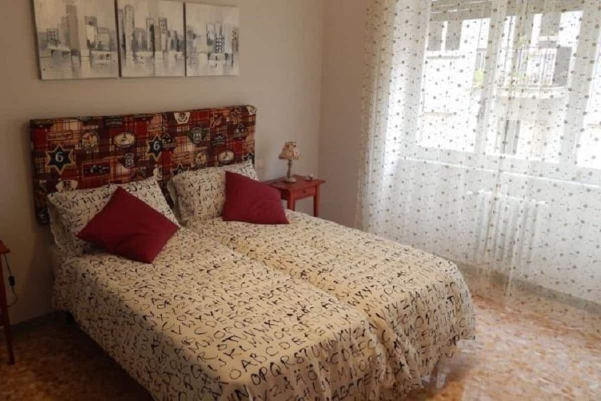 A bedroom with a bed, two nightstands, a cityscape artwork, and a window with sheer curtains.