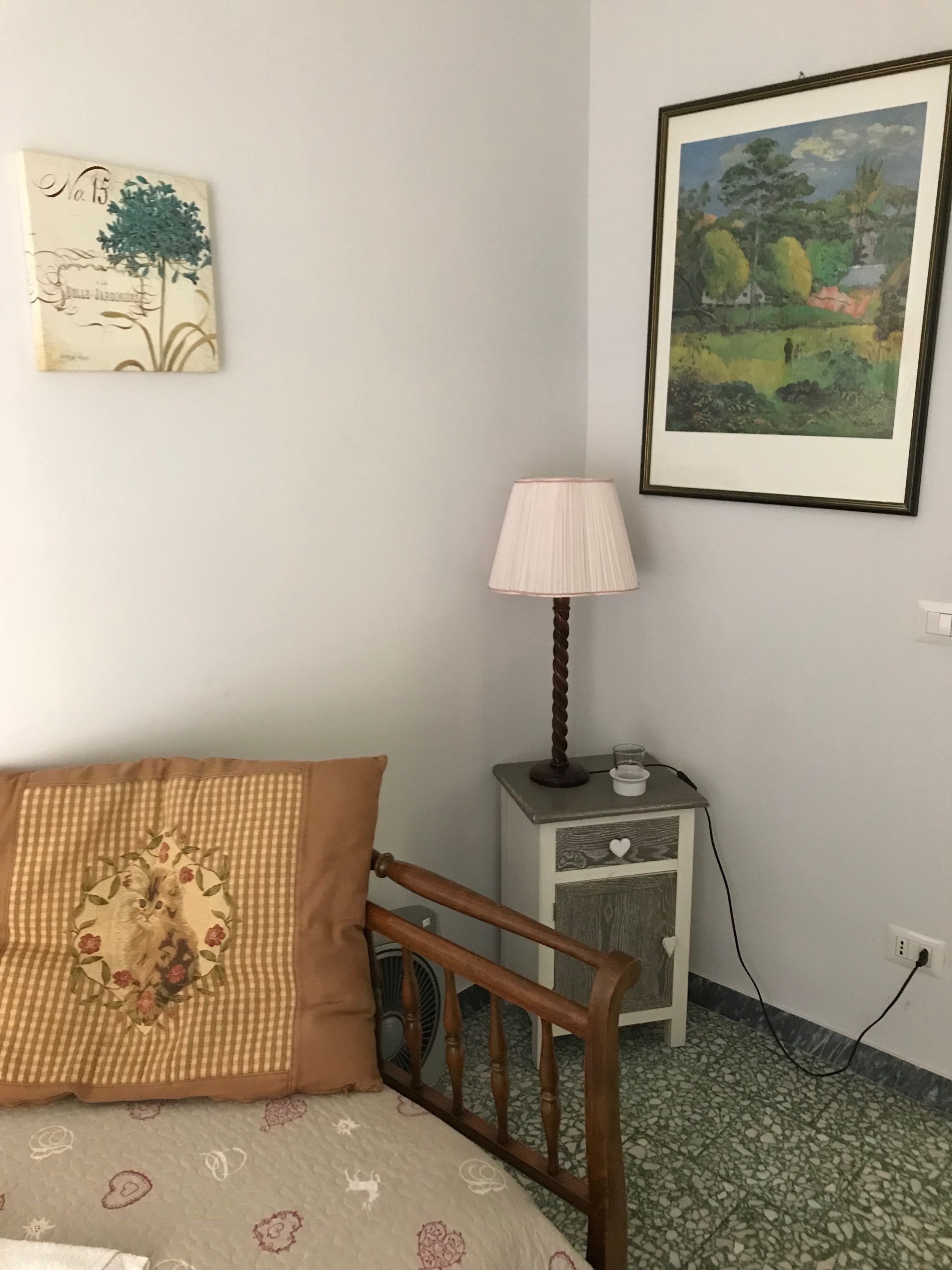 A bedroom with a bed, a bedside table with a lamp, a small cabinet, and two framed artworks on the wall.