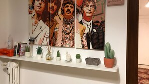 A wall with three framed artworks depicting a man in a suit, a shelf with various items including books, a cactus, and a small basket. - Musei Vaticani Rooms (Rome)