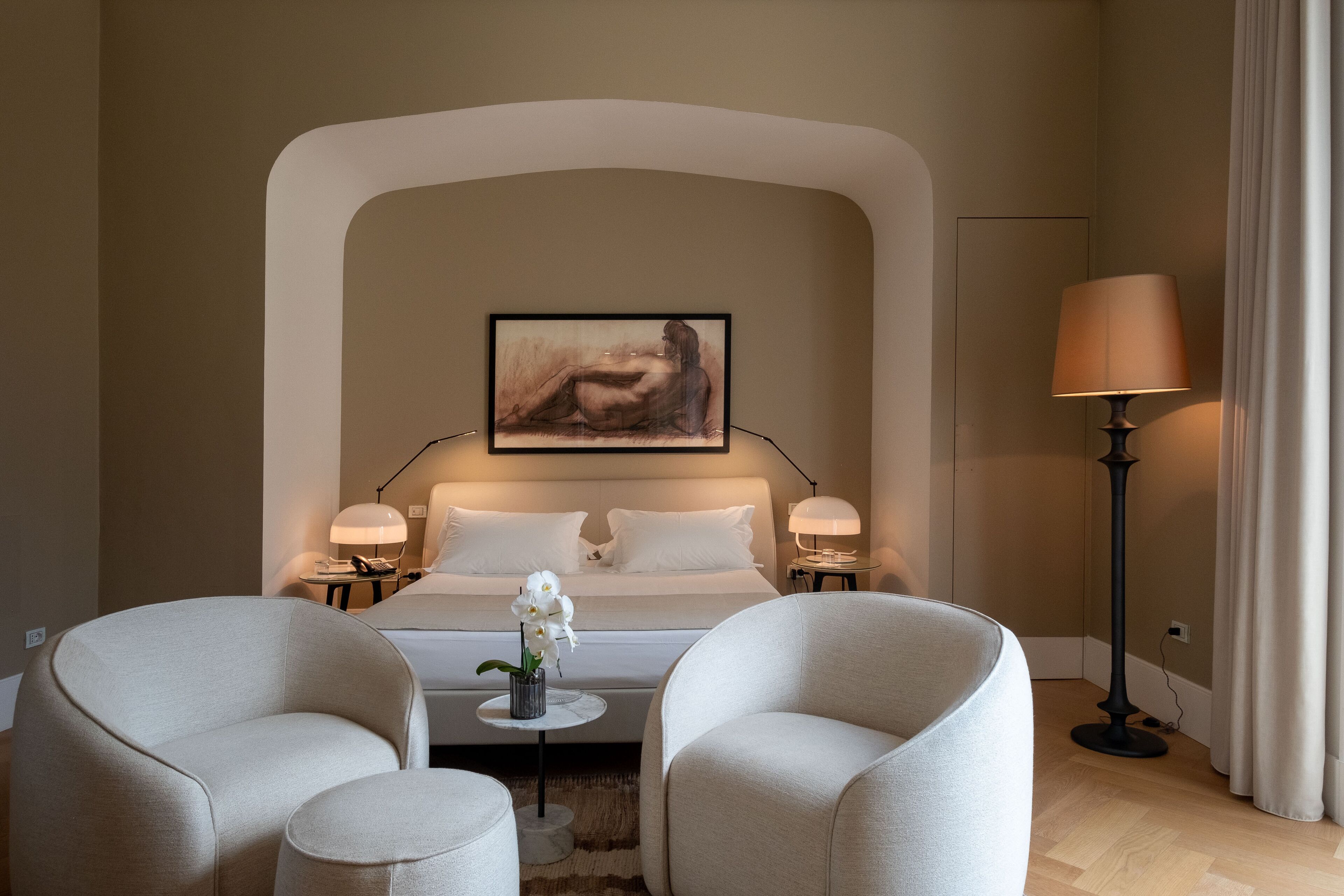Une chambre d’hôtel moderne avec un grand lit, deux tables de chevet avec des lampes, une petite table ronde ornée d’un vase de fleurs et deux fauteuils.
